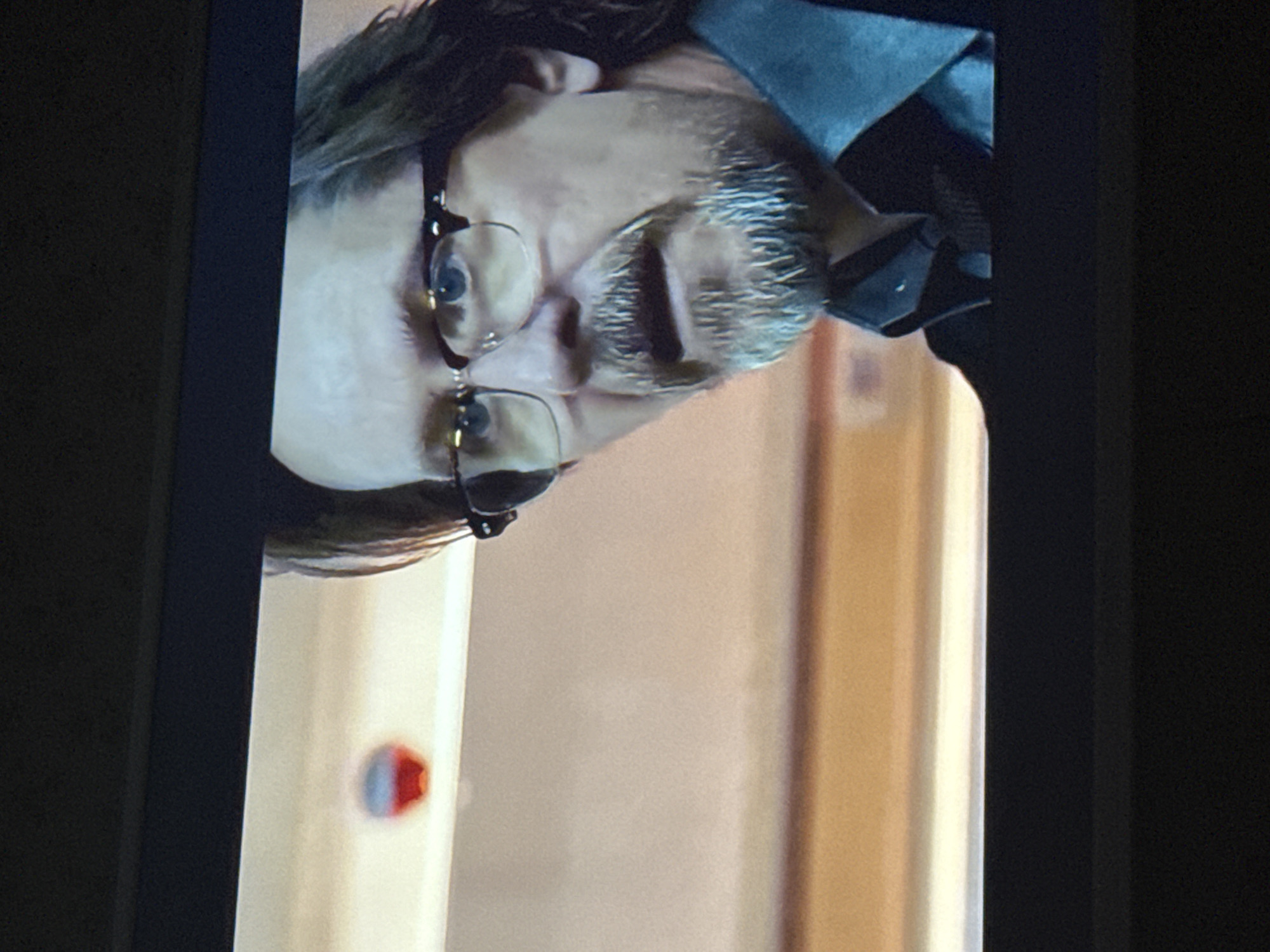 A man with glasses and a gray beard is shown in the image. He is wearing a blue collared shirt. The background appears to be an indoor setting, possibly a hallway, with beige walls and a door with a red element. The location is not discernible from the image.
