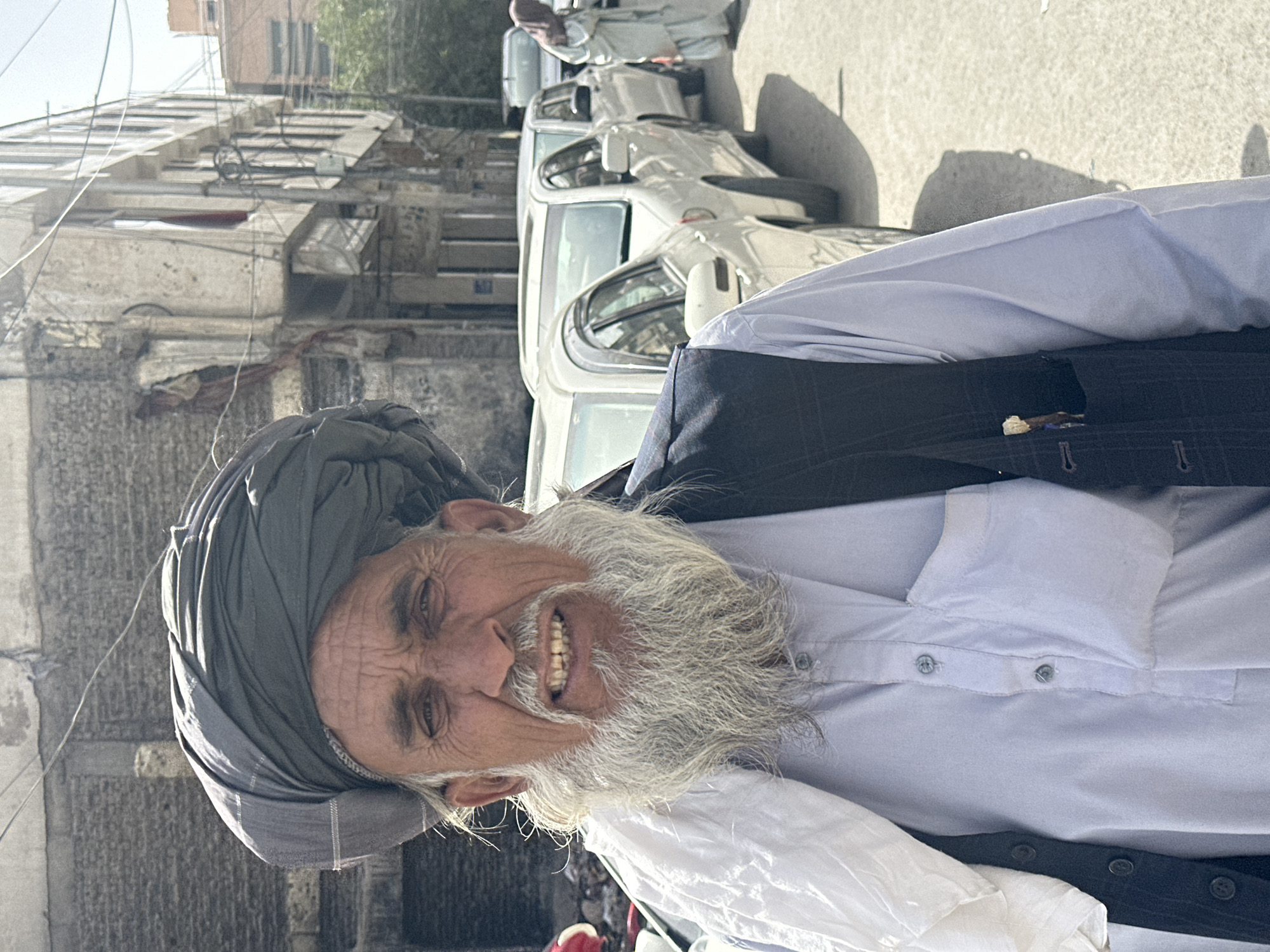 A man with a gray turban and a long white beard stands in the foreground, facing the camera directly. He is wearing a light blue shirt and a dark vest with checkered patterns. His shirt has buttons down the front and a pocket on the left side. A white cloth is draped over his left shoulder. His teeth are visible, suggesting he may be smiling or speaking.
Behind him, several white cars are parked in a line on a paved street. The cars appear to be modern sedans. In the background, a multi-story building with peeling paint and visible electrical wires stands on the left. Another building, possibly a different structure or part of the same complex, is visible further back on the right. The environment suggests an urban setting, consistent with Quetta City Tehsil, Pakistan. The light is bright, casting shadows on the ground, indicating it is daytime. A person in a light-colored garment and hijab is visible in the far right background, walking away from the camera. The overall scene captures a street view with a prominent individual as the central subject.