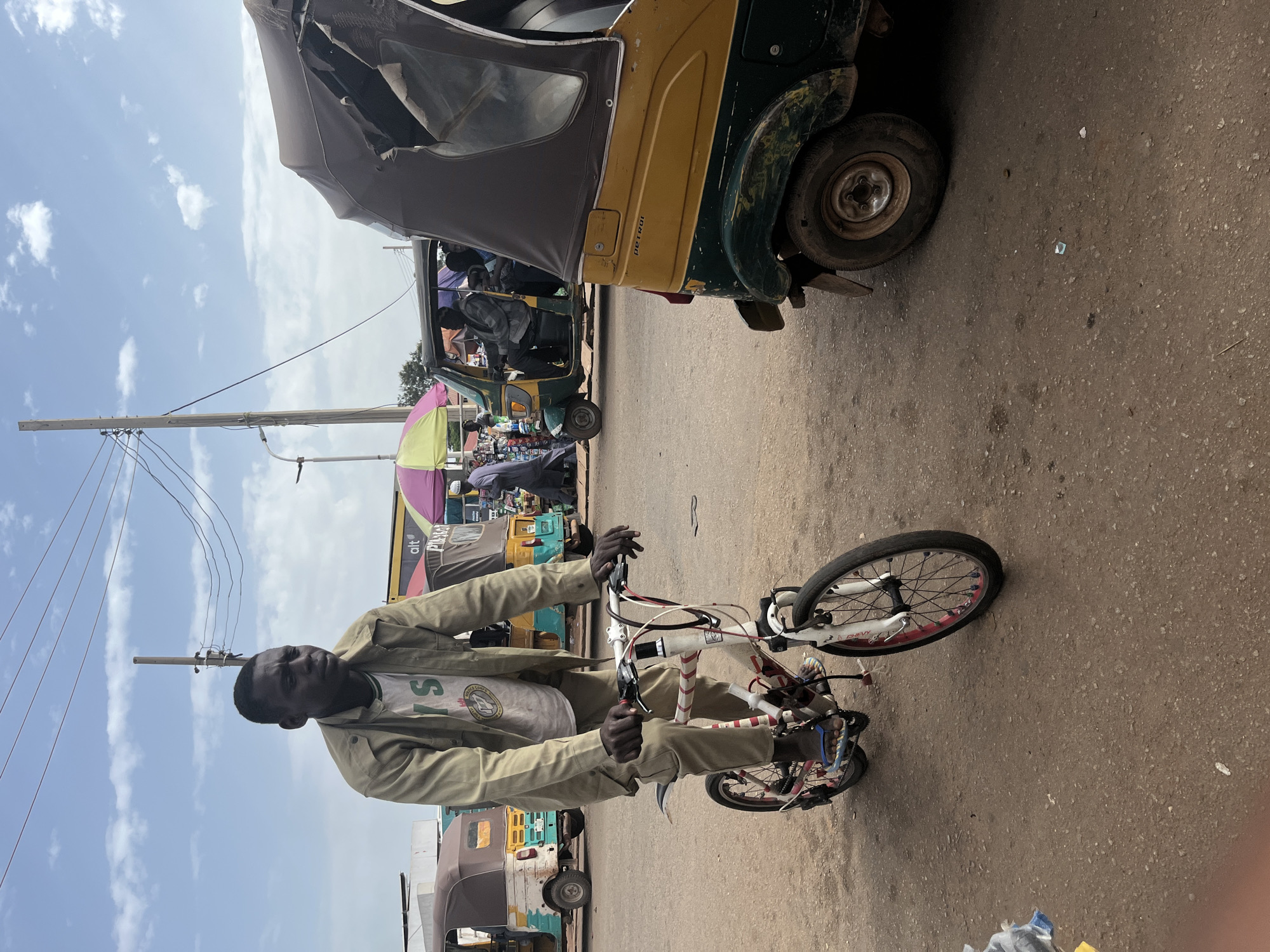 A man rides a bicycle down a dirt road in Gura Top, Nigeria. The man is wearing a tan jacket over a white t-shirt and tan pants. The bicycle is white with red accents. In the background, several auto rickshaws, also known as "keke napeps," are parked or driving. One auto rickshaw in the foreground is yellow and green. Another auto rickshaw in the background is white and green. A few people are visible in the auto rickshaws. Overhead, power lines are strung between utility poles. The sky is partly cloudy. The ground is a mix of dirt and small stones.