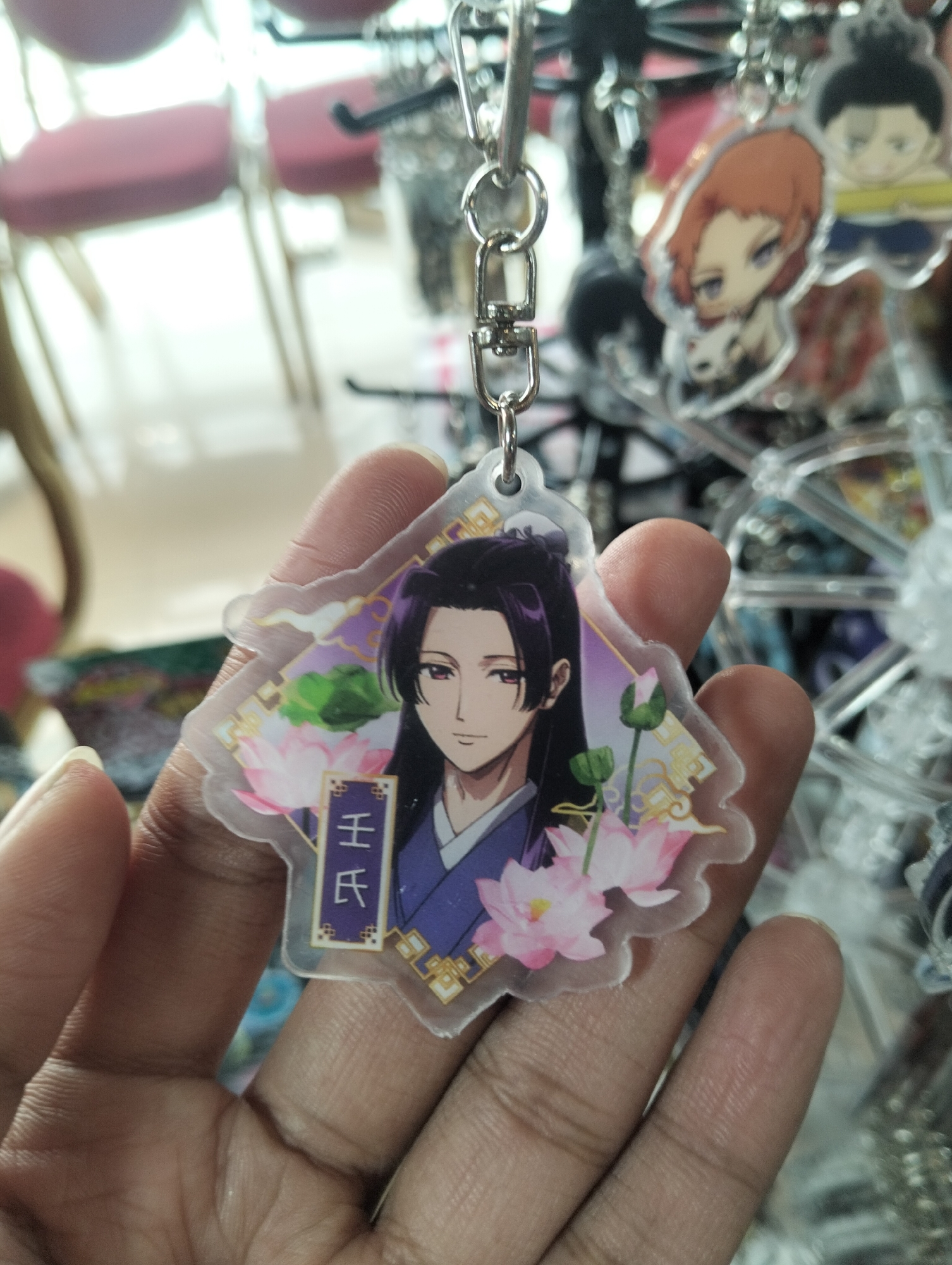 In the city of Dhaka, Bangladesh, a person's hand is holding a clear acrylic keychain. The keychain features an anime-style illustration of a male character with dark hair and purple clothing, surrounded by pink lotuses and decorative golden patterns. A small purple banner with Chinese characters is visible below the character's chest. The background is blurred, showing glimpses of other keychains and what appears to be furniture, suggesting the image was taken inside a shop or market.