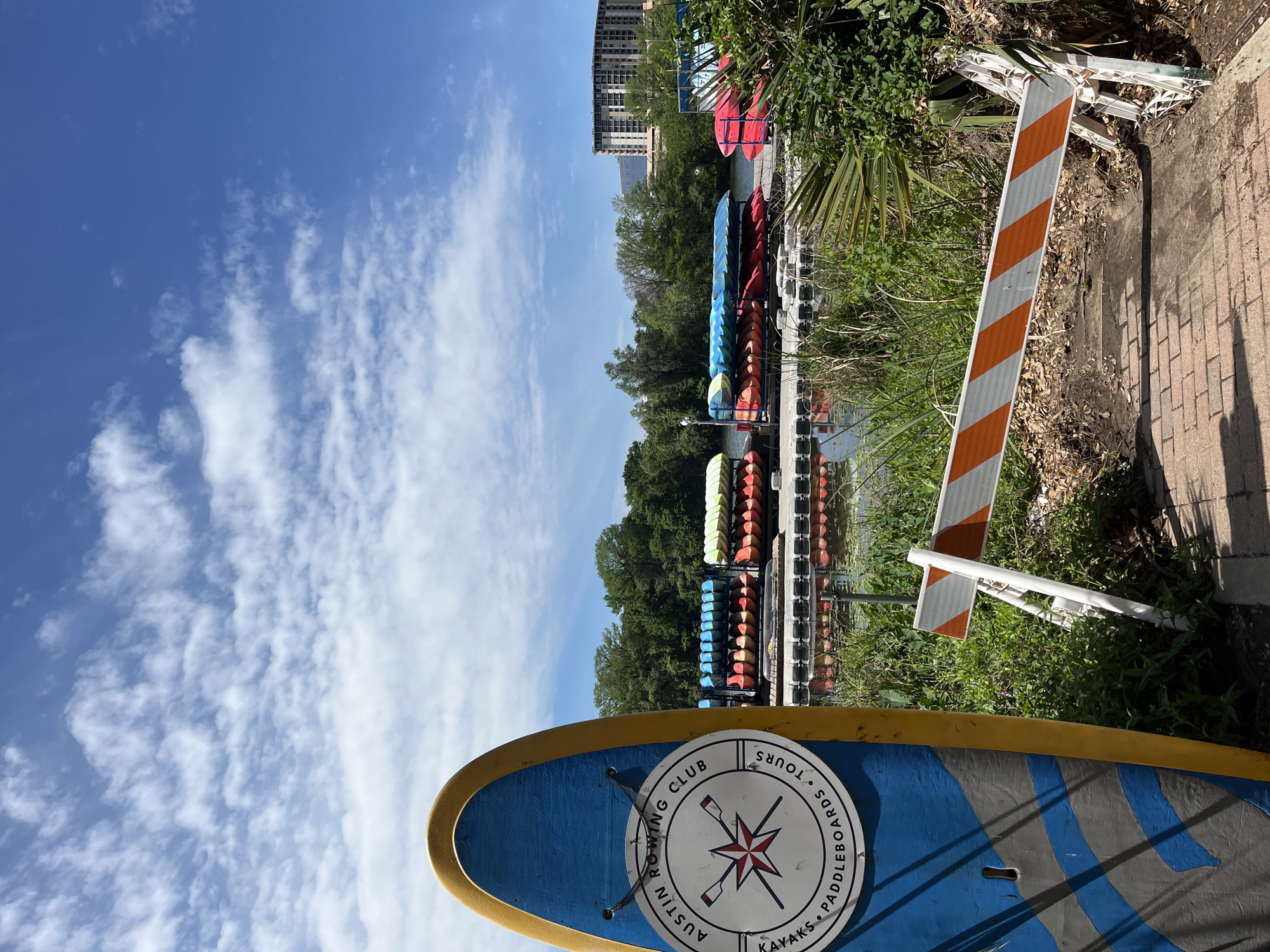 The photograph depicts a scenic view in Austin, Texas, USA, focusing on the Austin Rowing Club's location on Lady Bird Lake. A paddleboard with the club's logo, indicating the brand and function, lies in the foreground. The logo features the words "Austin Rowing Club, Kayaks, Paddleboards, Tours."
In the midground, a series of kayak racks are visible along the waterfront, filled with kayaks in various colors. Lush green foliage and trees separate the lake from the city skyline, which features modern high-rise buildings. The sky above is bright blue with scattered white clouds.
An orange and white striped barrier is situated between the paddleboard and the kayak racks, potentially indicating a hazard or access restriction. The image is taken during daylight, showcasing the natural and urban elements of the Austin cityscape.