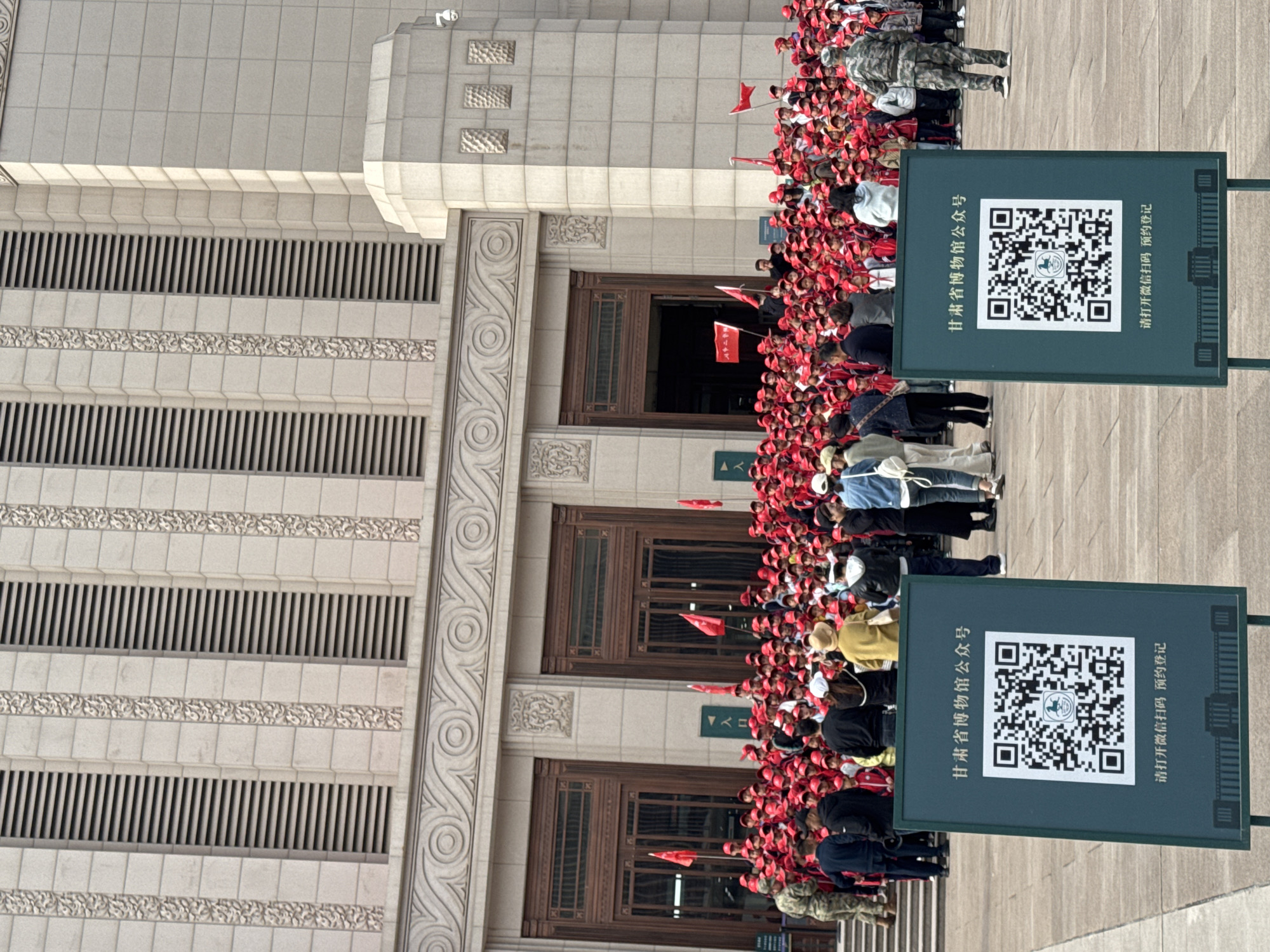 The image shows a large group of people, many wearing red hats and holding small red flags, gathered in front of a building. Two signs with QR codes stand in the foreground, labeled in Chinese as "Gansu Provincial Museum Public Account" and "Scan WeChat to make an appointment." The building has a stone facade with decorative architectural details and large wooden doors. The location appears to be the Gansu Provincial Museum based on the sign, which is situated in the Jianlan Road District of China.