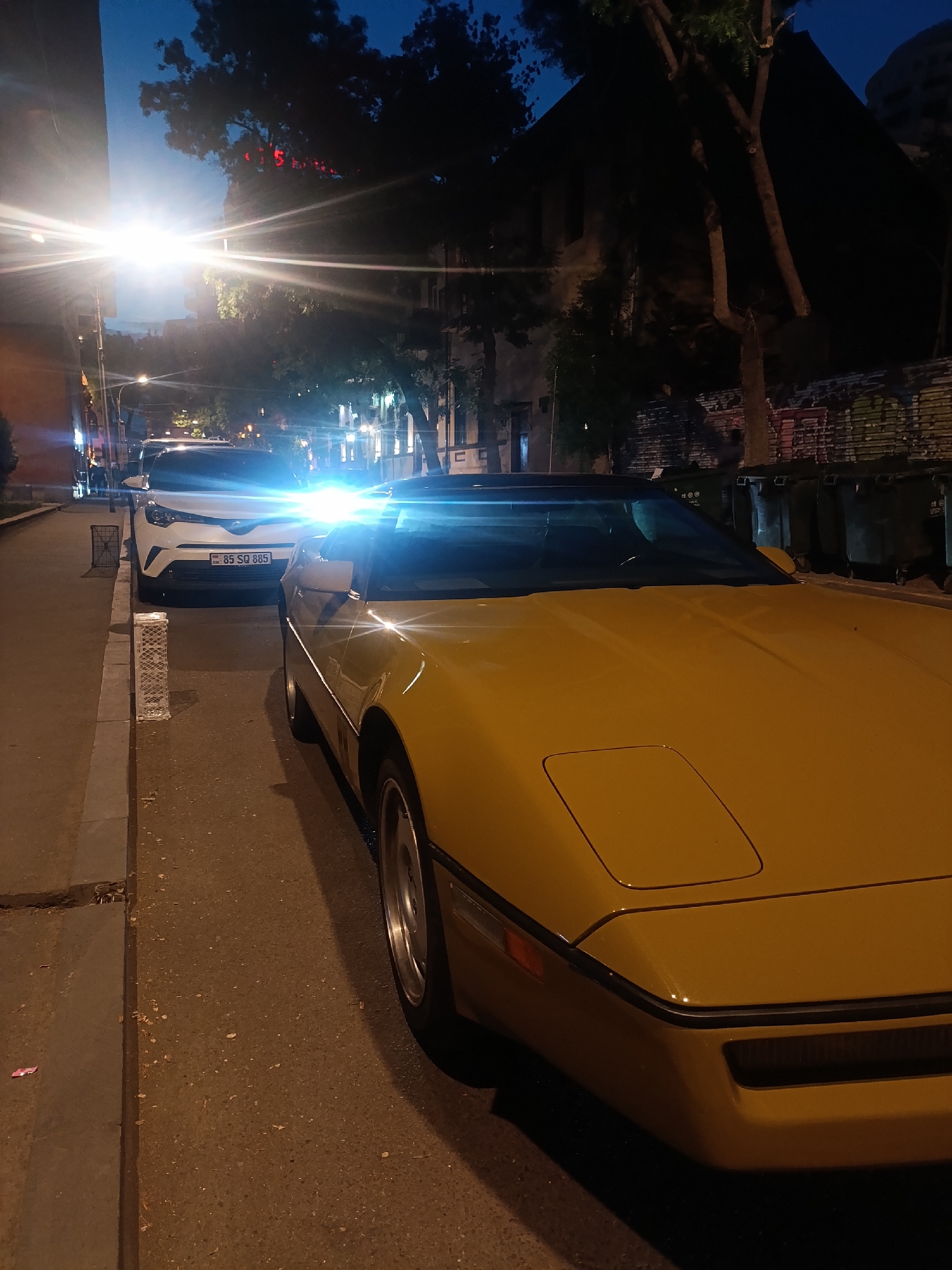The image depicts a street scene at night in Yerevan, Armenia. A yellow Corvette is parked along the street's edge, with a white Toyota C-HR parked behind it. The license plate of the Toyota reads "85 SQ 885". Streetlights cast strong glows throughout the image, obscuring details in the background. Trees and buildings are visible in the background, suggesting a residential or commercial area. Green trash containers are at the side of the building. The sky is dark blue, indicating it is either dusk or night. The street is paved and shows some wear. The angle of the photo is taken from the side of the Corvette, with the Toyota in the background.