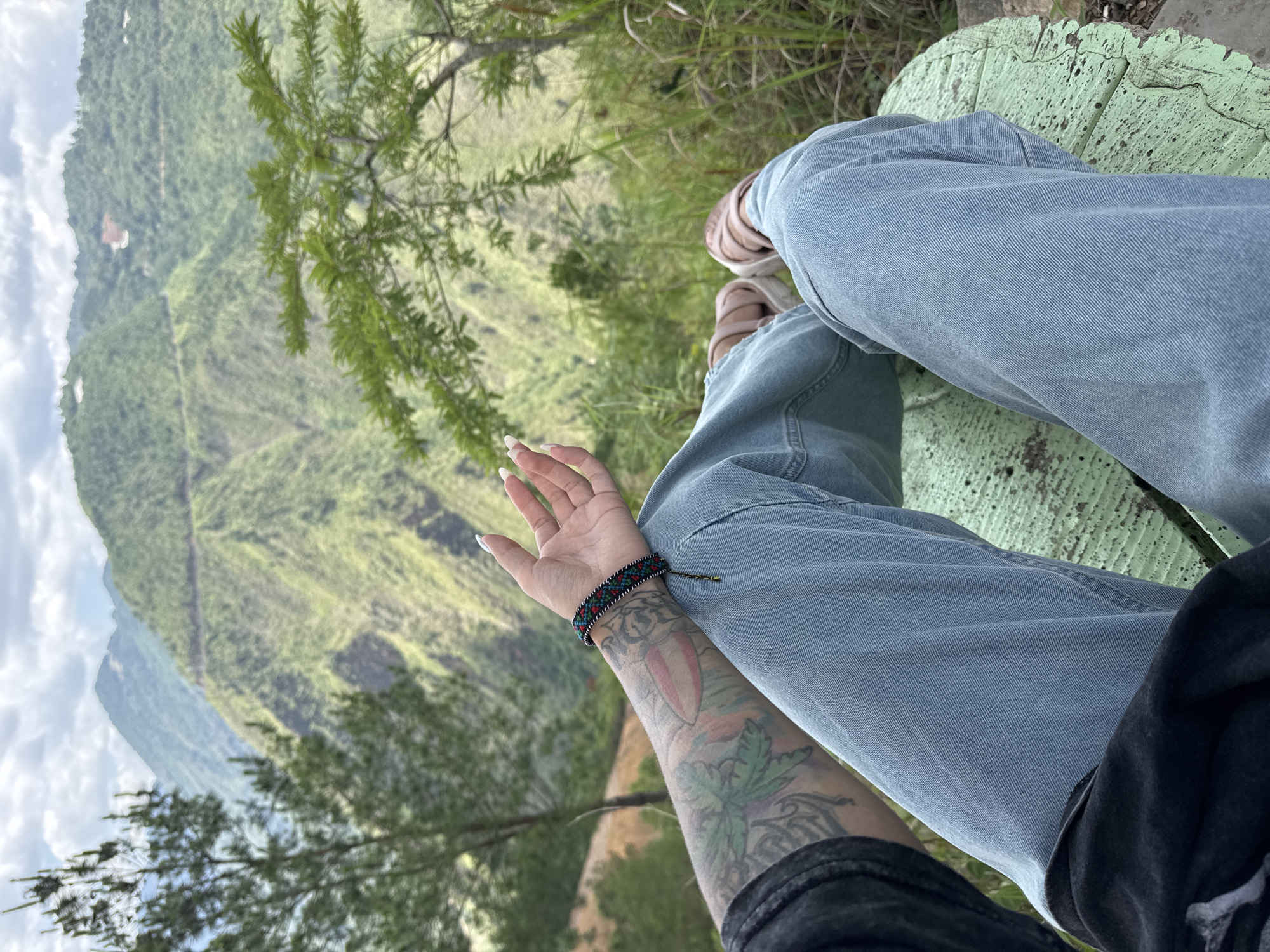 A first-person perspective shot shows a person seated outdoors in Lũng Cú Commune, Vietnam. The person wears light blue jeans and sandals, with an arm resting on the leg, revealing tattoos and a bracelet. A light green, weathered wooden seat is visible. The background features a mountainous landscape with lush green vegetation under a cloudy sky.