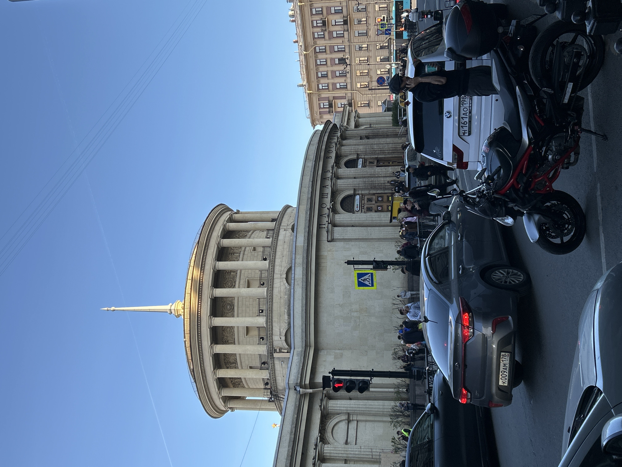 This image captures a vibrant street scene in Saint Petersburg, Russia, centered around a grand neoclassical building. The prominent structure is circular, adorned with numerous columns, and capped with a dome-like roof featuring a slender spire. Along its upper facade, the text "ПЛОЩАДЬ ВОССТАНИЕ" (Ploshchad Vosstaniya, or Uprising Square) is clearly visible, identifying the location.
The street below is bustling with activity. Several vehicles are visible, including a dark grey Kia sedan with the license plate "K009AM 178" and a white van displaying "H161AO 198." Both "178" and "198" are regional codes for Saint Petersburg, Russia. A black and red sport motorcycle is parked prominently near the white van. A red traffic light indicates that vehicles are currently stopped. A pedestrian crossing sign is also visible.
Numerous people are present on the wide sidewalks, some walking, others gathered in groups, or potentially seated at outdoor establishments along the building's base. The scene is illuminated by bright daylight under a clear blue sky, suggesting good weather conditions. The architectural style of the surrounding multi-story buildings, partially visible in the background, further contributes to the urban European setting.