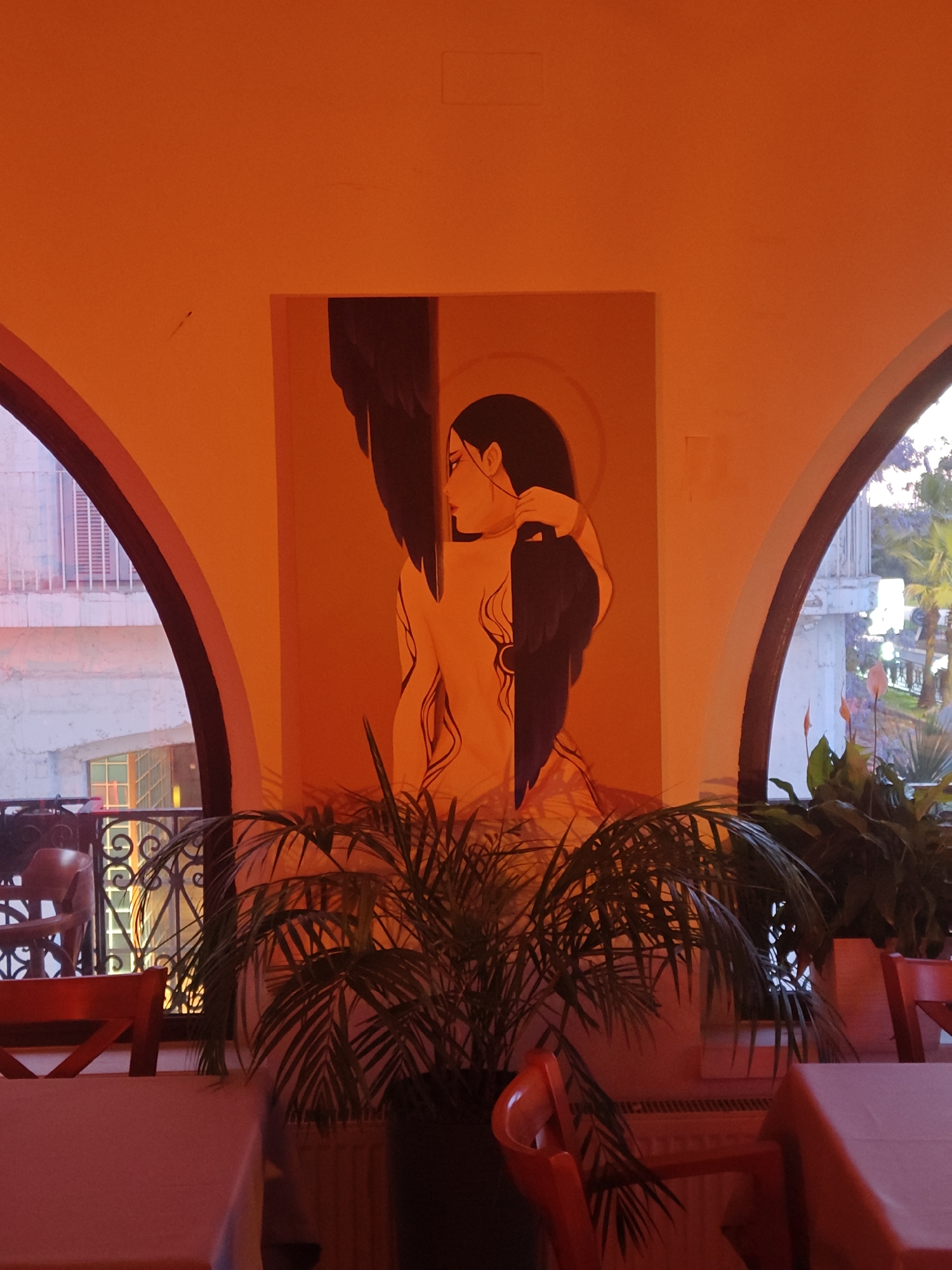 The image depicts an interior scene, likely a restaurant in Unknown, Unknown, characterized by its arch-shaped windows and warm orange lighting. A painting of a woman with dark hair and what appears to be wings is centered between two arched windows. A plant in a pot is positioned in front of the painting. Tables with tablecloths and red chairs are visible in the foreground, indicating a dining area. The exterior view through the windows includes buildings and trees.