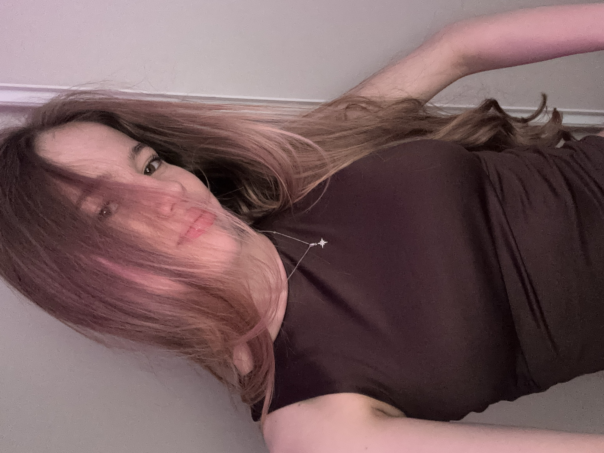 A young woman is captured in a close-up, rotated photograph, likely a selfie, taken indoors in Novoe Devyatkino, Russia. She is shown from the head to the upper torso, looking directly at the camera with a gentle, relaxed expression. Her long, wavy brown hair, featuring lighter, possibly pinkish-blonde highlights, flows across her face, partially obscuring one eye while the other is clearly visible with prominent eyelashes. Her lips are slightly parted in a subtle smile.
She is wearing a dark brown or black high-neck top or dress. A delicate silver necklace with a distinctive star or constellation-shaped pendant adorns her neck, resting on her collarbone. The background is a plain, light-colored wall, possibly off-white or light grey, with some areas exhibiting a soft pinkish hue, which could be due to lighting or photographic effects. The low camera angle suggests she might be lying down or holding the camera below her face. The lighting appears to be artificial or from an indoor setting without strong natural light, making the time of day unclear. No visible text is present in the image.