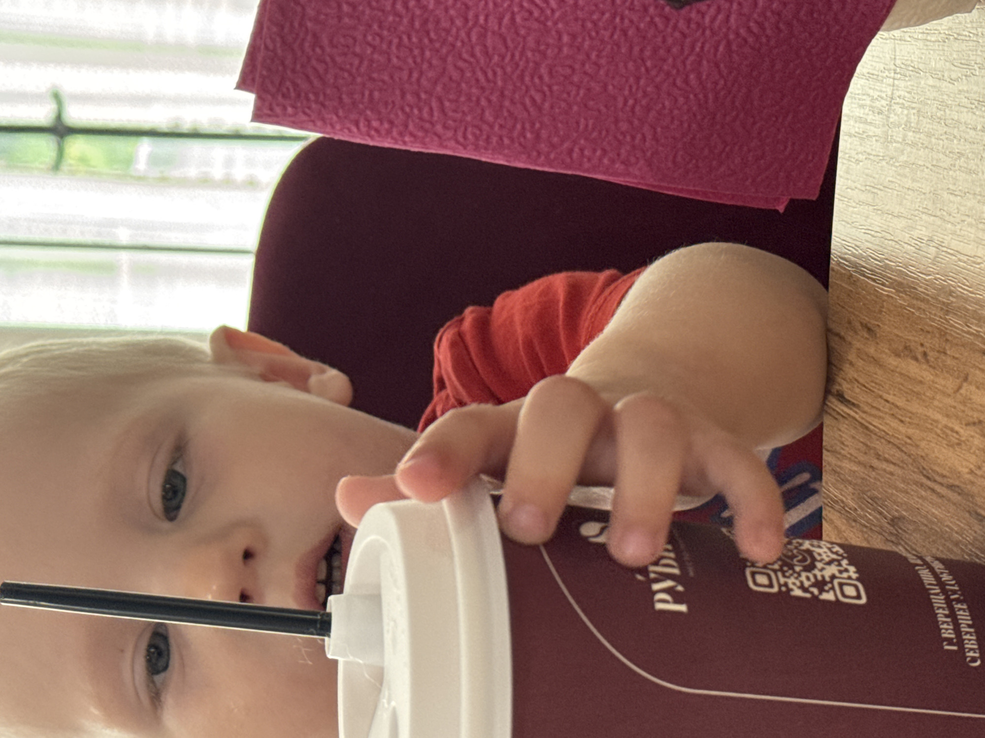A close-up, medium shot shows a fair-skinned toddler with light blonde hair reaching for a beverage cup with a black straw. The child wears a red t-shirt. A pink napkin is to the right of the child. The child's hands are visible as they grip the cup. The cup is maroon and features a white lid and straw. Text on the cup reads "РУБЛЬ" and a QR code. Below this, partially visible text states "Г.ВЕРЕЩАГИНО" and "СЕВЕРНЕЕ У.ЛОФТ". The background shows a dark red chair and a window with sheer curtains. The scene appears to be indoors, possibly in a cafe or restaurant in the city of Vereshchagino, Russia.