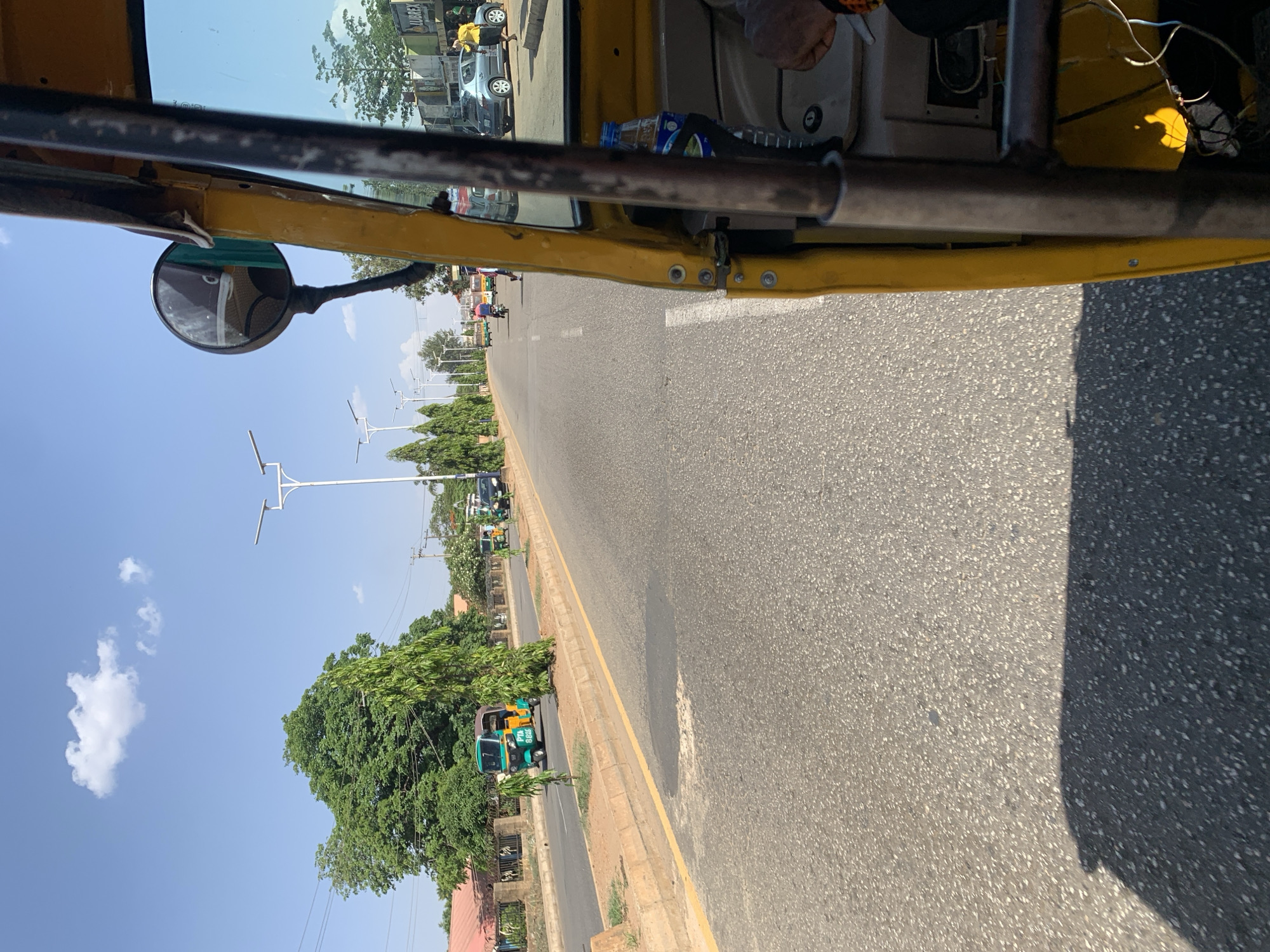 The image depicts a view from inside a yellow auto-rickshaw (keke) moving along a road. The foreground shows the interior of the keke, including metal framing and a side mirror. Through the windshield, a straight, paved road stretches into the distance under a blue sky with scattered clouds. On either side of the road are trees, streetlights, and buildings, including what appear to be houses with red roofs. Other kekes can be seen along the road. The location is New Stadium, Nigeria, as evidenced by the typical infrastructure and keke usage common in Nigerian cities. There are no visible people actively interacting in the street scene.
