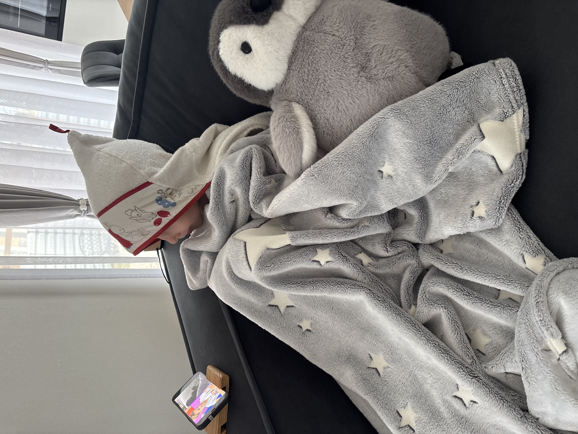 A young child is resting on a dark-colored sofa, covered with a gray blanket decorated with white stars and a white towel hood. A plush penguin toy is beside the child. A smartphone is positioned near the child, displaying a video. The scene is indoors, near a window with white blinds. The location is not specified as Chaikovsky, Russia, but that information may be inferred from the context in which the image was provided.
