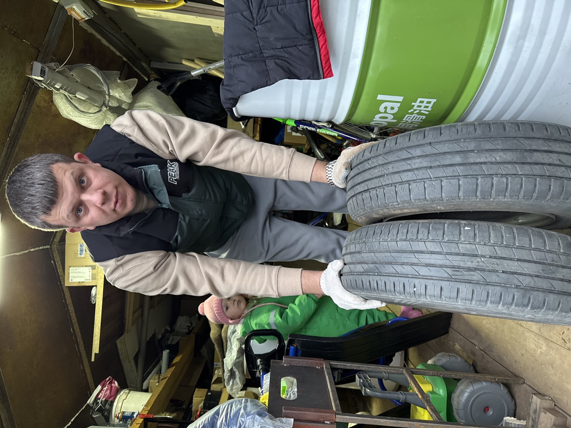 An adult male and a child are present within a cluttered indoor storage space, likely a garage or workshop, in Gornyi (Dvurechenskoe S/P), Russia. The male, positioned centrally in the foreground, possesses short dark hair, blue eyes, and wears a dark green puffer vest over a light beige long-sleeved shirt and grey sweatpants. He looks directly at the camera with a neutral expression, his hands, covered in white work gloves, supporting two upright car tires on a wooden floor. A metallic watch is visible on his left wrist. To his left and slightly behind, a young child wearing a pink knit beanie and a bright green full-body suit stands, looking towards the left of the frame. The environment features wooden plank walls and ceiling, densely packed with various items. On the left, shelves hold white buckets, plastic bags, and other containers. To the right, a large green and white industrial barrel, labeled "opal" and featuring Chinese characters for "lubricating oil," stands with a black puffer jacket draped over its top. Further objects include a green toy cart, cardboard boxes, exposed wiring, and a ceiling-mounted fan.