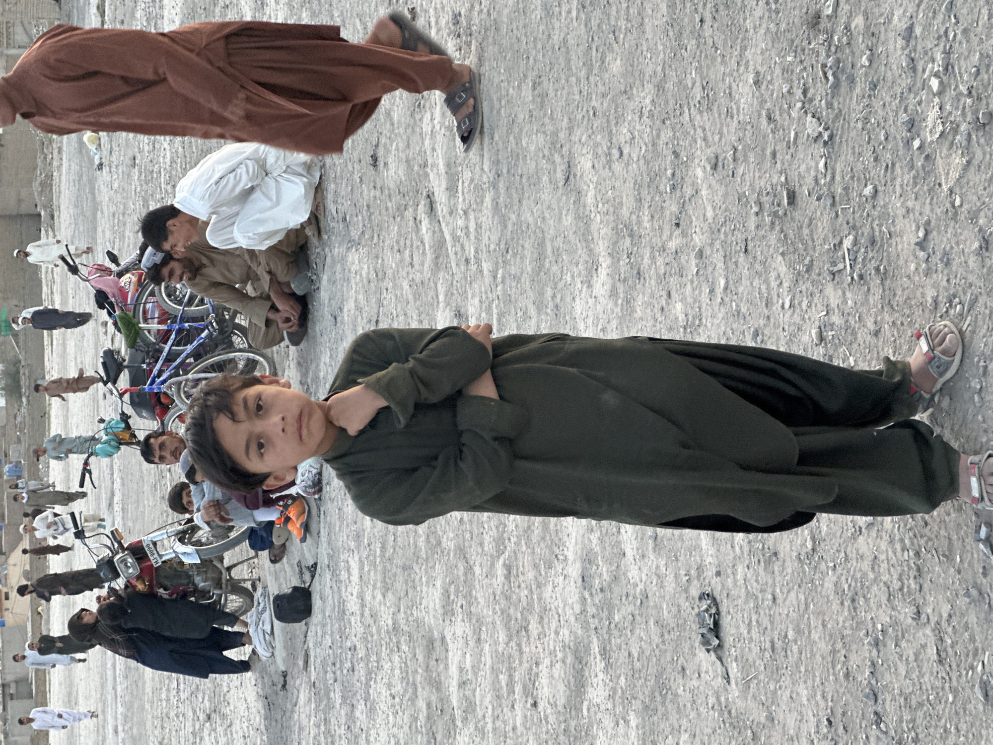 The photograph depicts a group of people in what appears to be a public space in Quetta City Tehsil, Pakistan. The ground is dirt and gravel. A young boy stands in the foreground, facing the camera with his arms crossed. He is wearing a dark green outfit and sandals. Behind him, there are several other people, including some sitting on the ground and some near bicycles and a motorcycle. Some people are wearing traditional Pakistani clothing. The overall scene suggests an outdoor gathering or casual meeting.