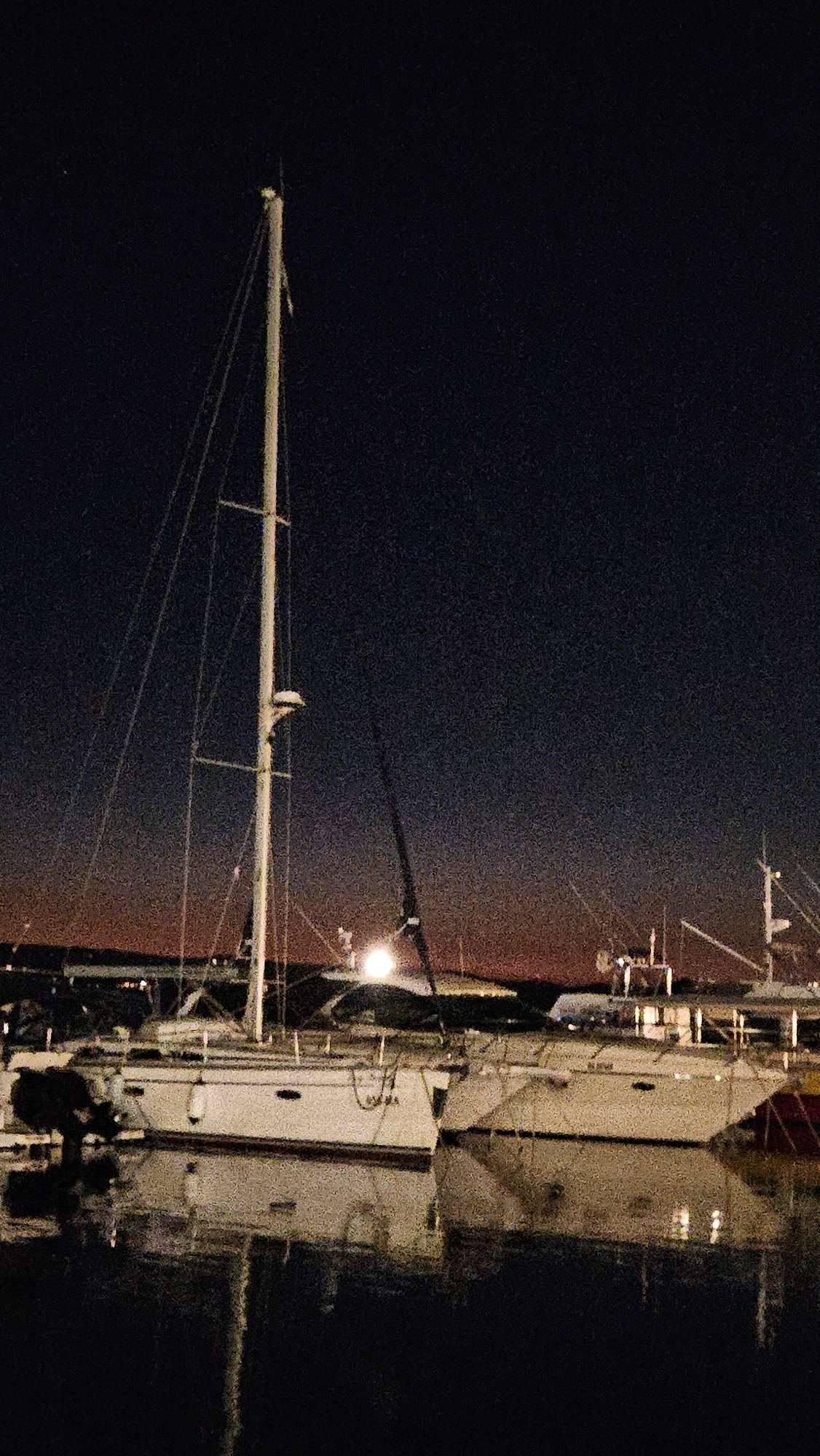 At night, in the harbor of Općina Tučepi, Croatia, a sailboat with a tall mast is docked. The sailboat is white with red trim and is reflected in the dark water. Other boats are also docked in the harbor, and the distant shore is visible with lights. The sky is dark with a hint of orange near the horizon. The scene is quiet and still.