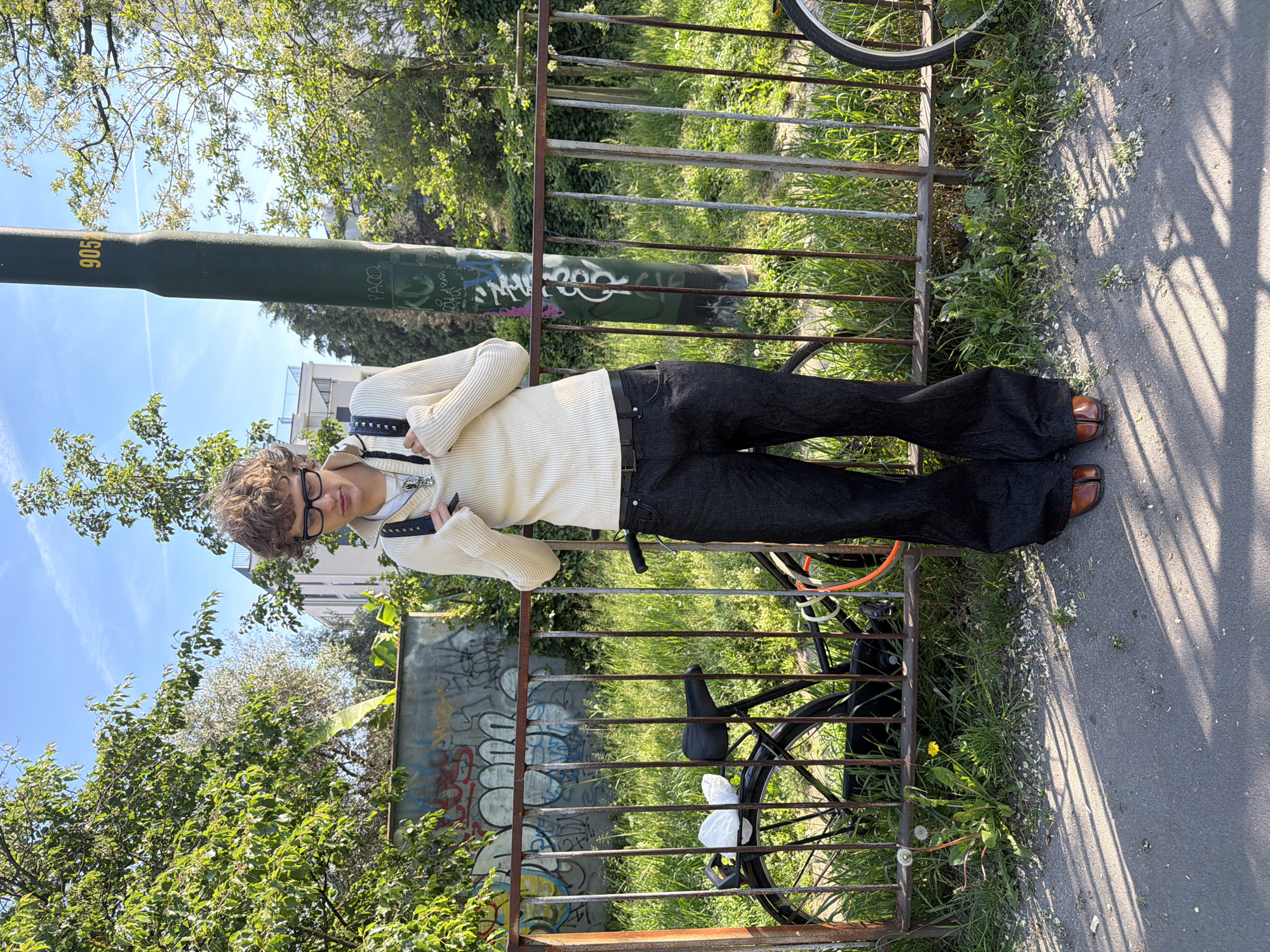 An outdoor, full-length portrait of a person standing beside a rusty metal fence, captured in daylight and rotated 90 degrees counter-clockwise. The subject, a young person with short, curly brown hair and black-rimmed glasses, is wearing a cream-colored ribbed long-sleeve top, dark wide-leg pants, and brown split-toe (tabi) shoes. A black belt with silver studs and a silver chain are visible accessories. The person stands casually with arms slightly bent, hands held in front of the body, and looks towards the camera.
Behind the subject, a tall, dark green cylindrical pole displays the yellow numbers "905" and white graffiti, including the word "PACO" and other markings. Beyond the fence is an unkempt grassy area where a dark-colored bicycle is visible, with a white plastic bag tied to its pedal or frame. Further back, lush green trees and bushes fill the scene, with some distant buildings visible through the foliage and a clear blue sky with a few clouds overhead. A paved asphalt path is in the foreground, marked by distinct shadows cast by the fence, indicating bright, sunny weather. A section of a concrete wall behind the fence is also covered in extensive graffiti. The scene suggests an urban green space, likely in Milan, Italy.