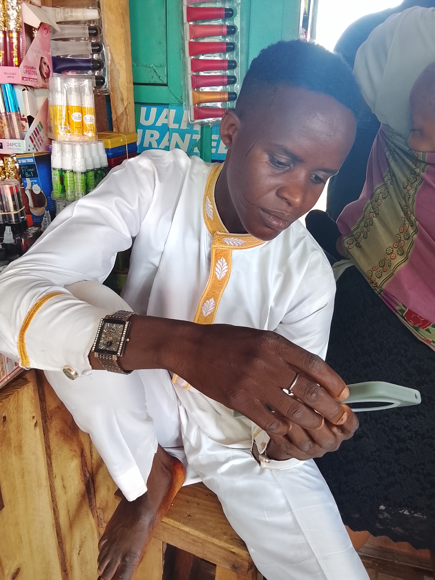 In the city of Akwanga, Nigeria, a young African man dressed in a white garment with gold trim is seated on a wooden bench. He is wearing a diamond-encrusted watch on his left wrist and a silver ring on his right hand. The man is looking down at a light green object he is holding, possibly a phone case or a small electronic device. Behind him, a woman in a pink and green patterned cloth is partially visible, carrying a baby on her back. The background suggests a market stall, with shelves filled with cosmetics and other products. The lighting is bright, indicating an outdoor or well-lit indoor setting.