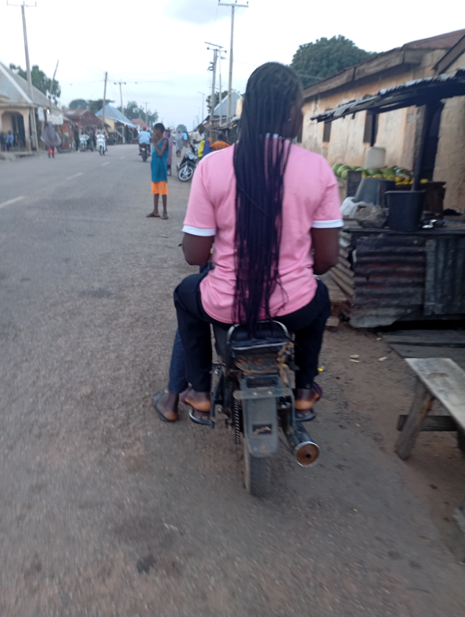 A person with long, dark braided hair is sitting on a motorcycle, facing away from the camera. They are wearing a pink t-shirt with a white collar and black pants. Their feet, clad in brown sandals, are resting on the motorcycle's footpegs. The motorcycle is parked on an asphalt road in what appears to be an urban environment. In the background, there are buildings with corrugated metal roofs and a fruit stand with green and yellow fruits. Other people and motorcycles are visible further down the road, suggesting a busy street. The sky is overcast. The scene is likely located in Shendam, Nigeria, based on the context provided.