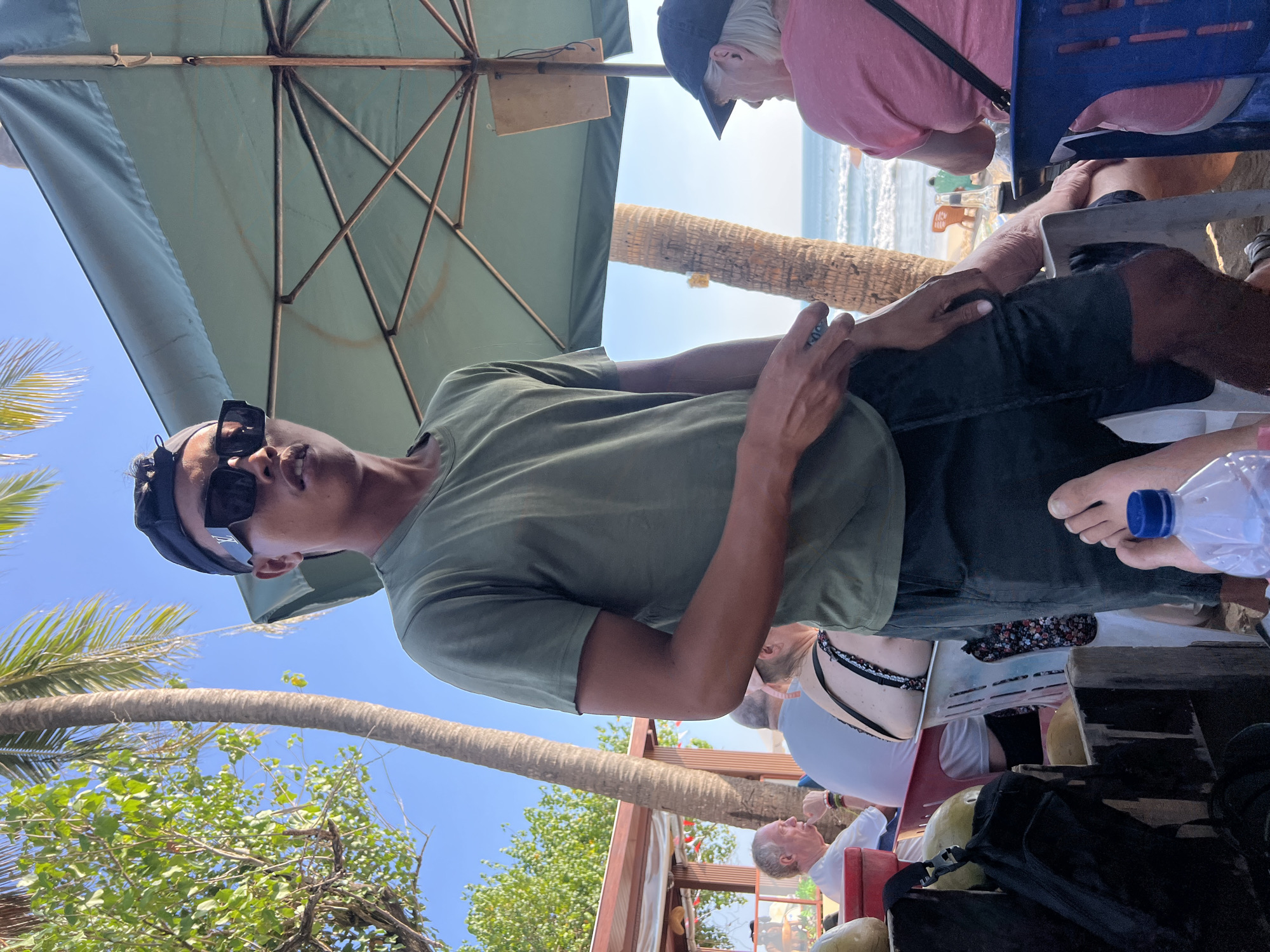 A man stands under a green beach umbrella in Kuta, Indonesia, wearing a dark baseball cap, black sunglasses, a green t-shirt, and dark shorts. He is looking towards the camera, with his left hand resting on his right leg. To his right, a woman with blonde hair and a pink shirt is seated, facing away from the camera. In the background, the ocean and a sandy beach are visible, with palm trees and other beachgoers scattered around. The overall scene depicts a casual day at the beach in Kuta.