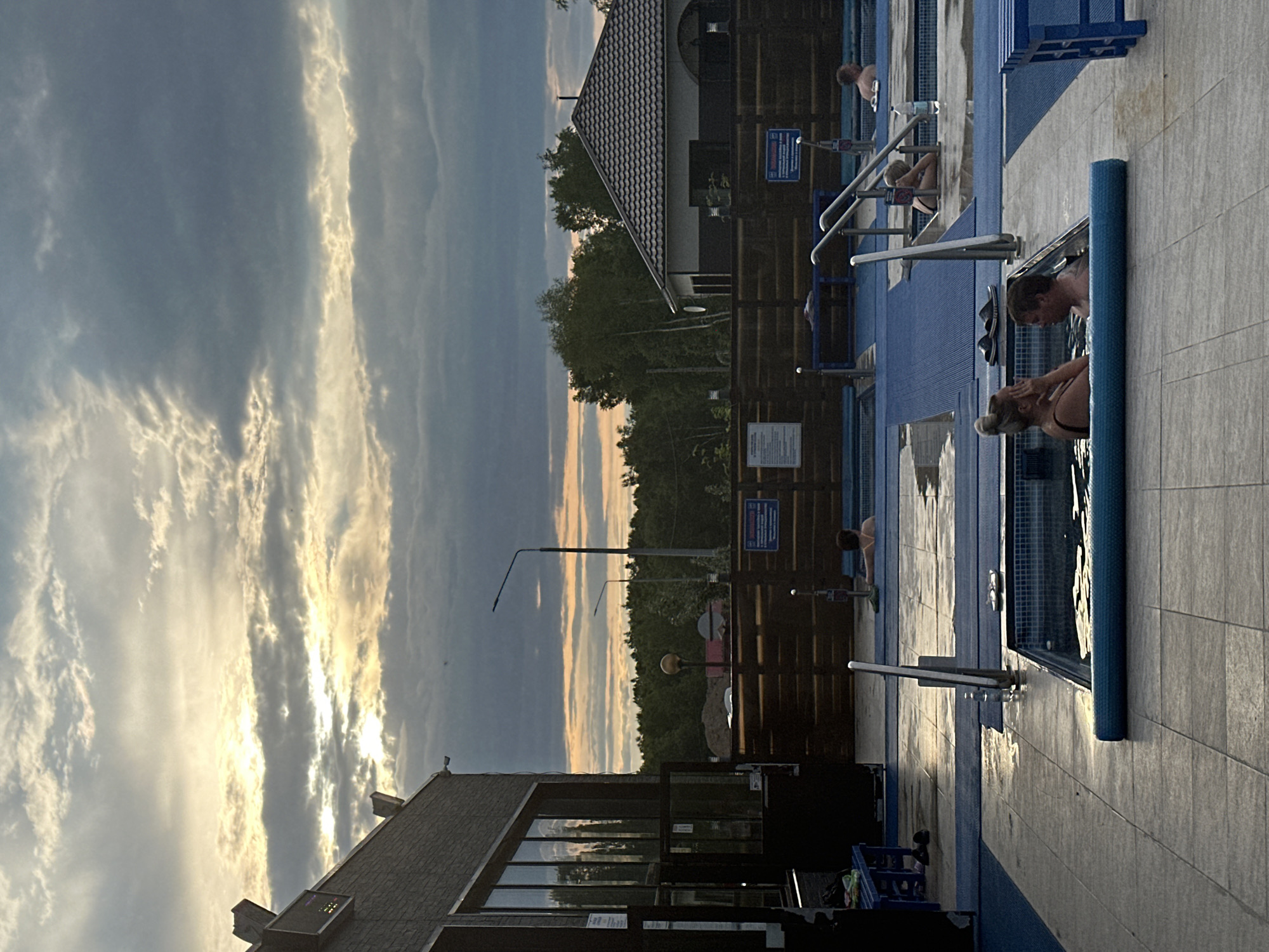 The image depicts an outdoor hot tub facility at dusk. The sky is filled with dramatic, cloudy formations, with rays of sunlight breaking through, casting a warm glow. In the foreground, two individuals, a woman with her hair in a bun and a man, are relaxing in a hot tub. The woman is touching her face, while the man is looking towards her. They are both wearing swimwear.
Further back, other people are visible in separate hot tubs. One woman is leaning back in a hot tub, looking upwards. Another person, partially visible, is also in a hot tub in the distance. The hot tubs are rectangular with tiled edges and are surrounded by blue mats and paved walkways. Metal railings and steps provide access to the tubs.
To the left of the image, a modern building with large windows is visible, suggesting it might be part of the facility's amenities. A wooden fence runs along the perimeter of the area, with trees and a building with a tiled roof visible beyond it. Streetlights are also present in the background. The overall atmosphere is one of relaxation and leisure, set against the backdrop of a fading sky. There is no indication in the image that this location is in Yuzhnyye Koryaki, Russia.