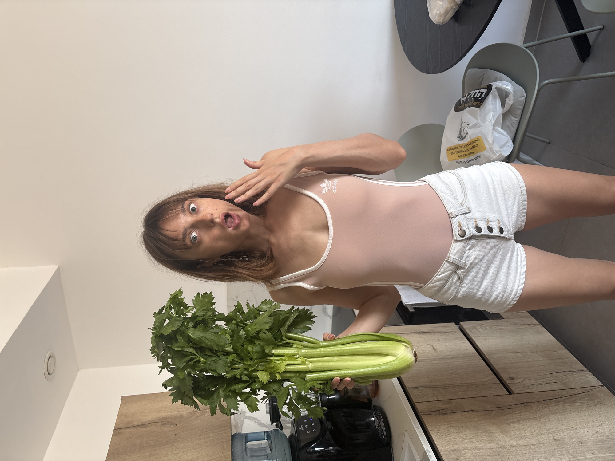 A young woman with brown hair and bangs stands in a kitchen, holding a large bunch of celery. She is wearing a pale pink one-piece swimsuit with white trim and an Adidas logo, and white denim shorts. Her expression is surprised, with her mouth open and one hand raised to her face. The kitchen appears modern, with wooden cabinets and white countertops. A water dispenser is visible on the counter. In the background, there is a dark round table with a chair that has a plastic bag on it. The lighting suggests it is daytime. Based on the visible text on the plastic bag, which includes Hebrew writing and potentially a phone number, the location is likely Tel-Aviv, Israel.
