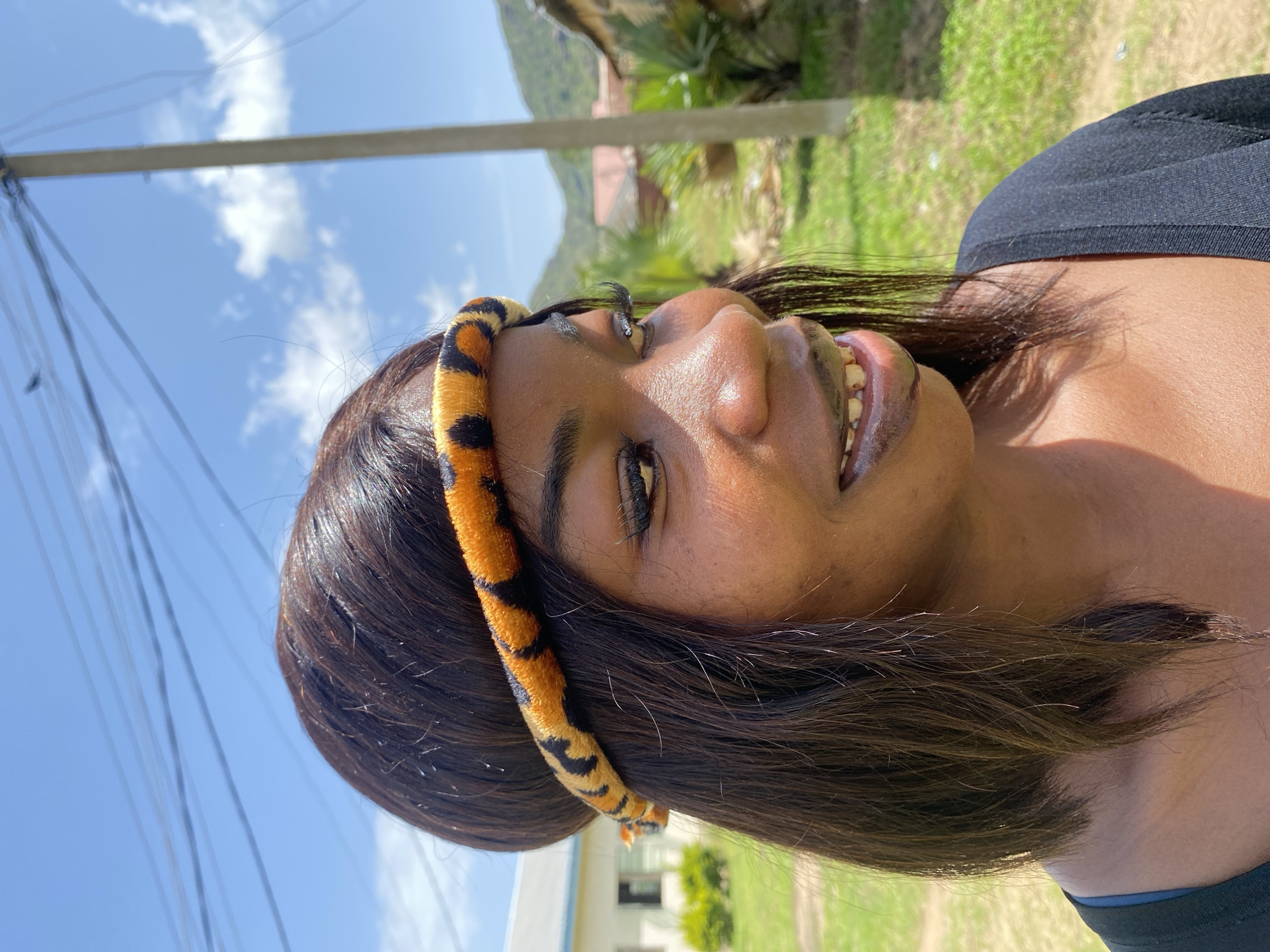 This is a close-up, vertically oriented, outdoor portrait of a young Black woman. She is wearing a black top and a fuzzy headband with a black and orange tiger-stripe pattern. Her dark hair is long and straight. Her head is tilted, and she is looking to her right, with a slight smile revealing her teeth. Her eyes are open, and her eyebrows are well-defined. The lighting suggests it is daytime, likely sunny, with bright highlights on her skin.
The background is somewhat blurred but includes greenery and what appears to be a building in the distance. There are also power lines and a pole visible in the upper portion of the image, indicating an outdoor, possibly semi-urban or rural, setting. The sky is blue with scattered white clouds. The overall mood is relaxed and candid. The image was taken in Wang, Nigeria. There is no visible text in the image.