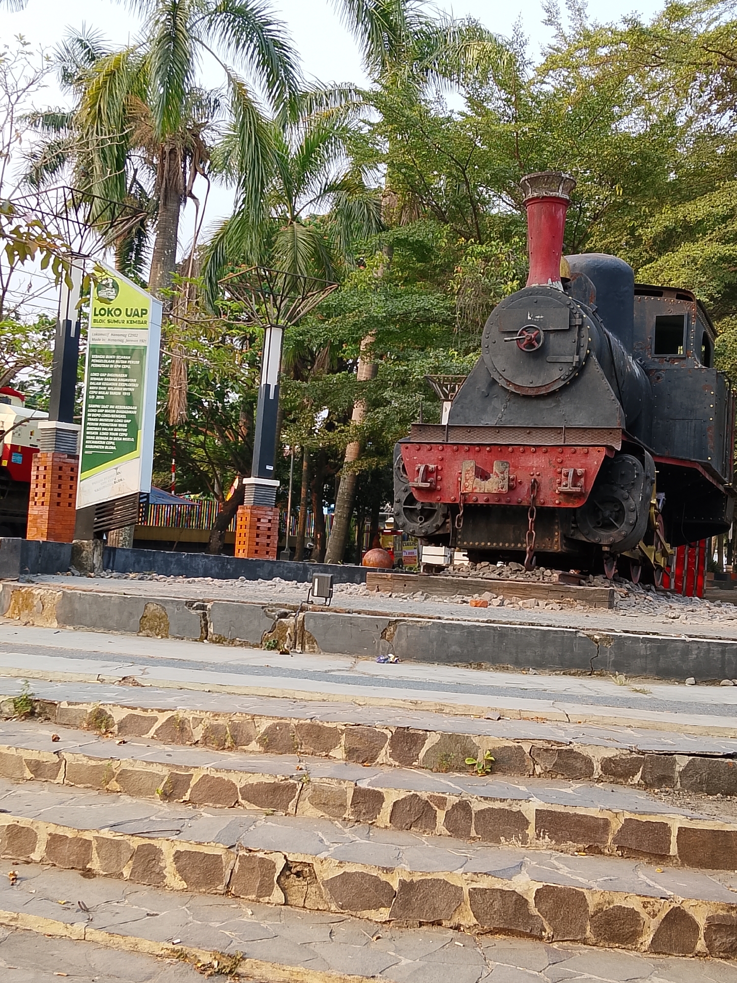 The image depicts an outdoor scene in Balun, Indonesia, focusing on a vintage black and red steam locomotive displayed on a track. The locomotive, which appears to be a preserved historical artifact, is positioned on a raised platform made of stone and gravel, situated in a park-like setting. In the background, lush green trees and tall palm trees create a tropical ambiance. To the left of the locomotive, a tall signpost with text and graphics provides information about the "LOKO UAP" (Steam Locomotive). A set of stone stairs leads up towards the viewing area in the foreground. The overall scene suggests a public space dedicated to historical preservation and tourism. There are no people visible in the image.