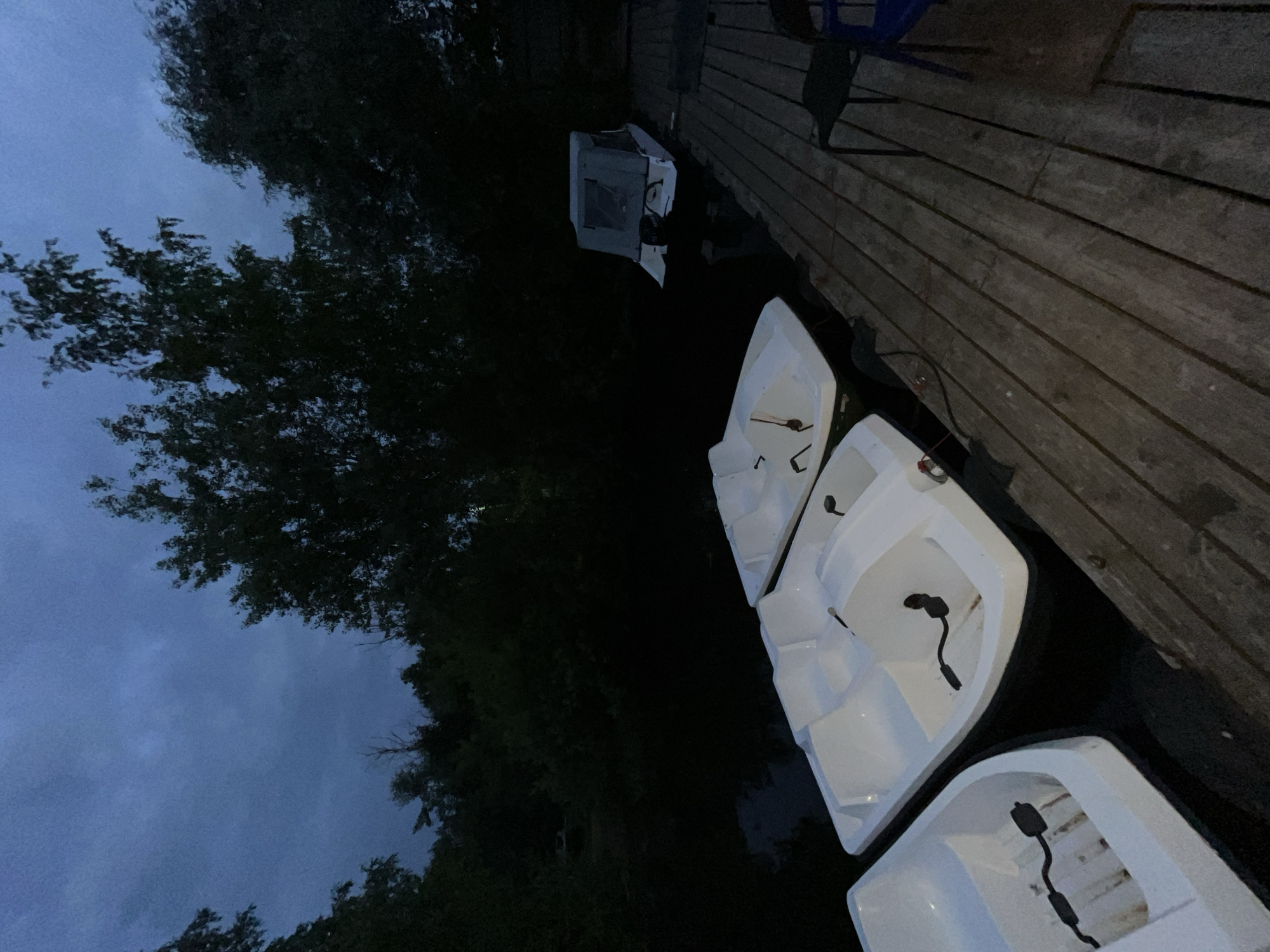The image depicts a twilight scene at a dock on the Kama River in Chaikovsky, Russia. Several white pedal boats are moored to the wooden dock, which extends into the dark water. In the background, dense trees line the riverbank, and the sky is a deep blue, suggesting the end of daylight. A small boat with a canvas cover is also visible in the water. The scene is quiet and serene, with no people present. The lighting is low, characteristic of dusk.