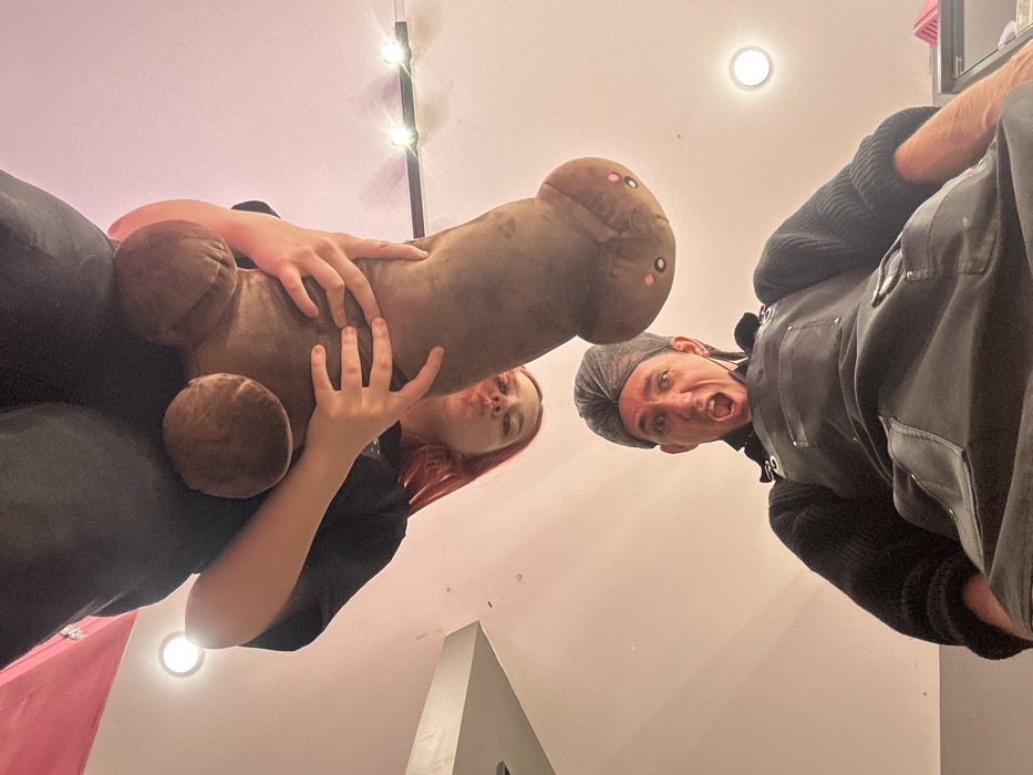 The image, taken from a low-angle perspective looking upward, depicts two individuals in an indoor setting. The specific location within Saratov, Russia, is not visually identifiable.
In the upper portion of the frame, a person with reddish-brown hair, wearing a dark shirt and dark pants, is positioned inverted. This individual holds a large, brown, phallic-shaped plush toy with both hands. The toy features two small black eyes and pink cheek dots. The person exhibits a pouting facial expression, directed towards the viewer.
Below this figure, a male subject is visible, wearing a dark grey headwrap, a black long-sleeved garment, and a black apron with multiple pockets and silver metallic hardware. His face displays an exaggerated expression, with his mouth wide open and eyes wide, looking directly upwards towards the camera and the plush toy.
The immediate background consists of a pale pink or light-colored ceiling and upper wall sections. Multiple light sources are present, including a circular recessed light fixture on the upper left, a linear strip of three smaller circular lights along a dark bar on the upper right, and another circular recessed light in the lower right. A grey architectural element is partially visible on the far left. A dark surface with a pink object rests in the lower right corner.