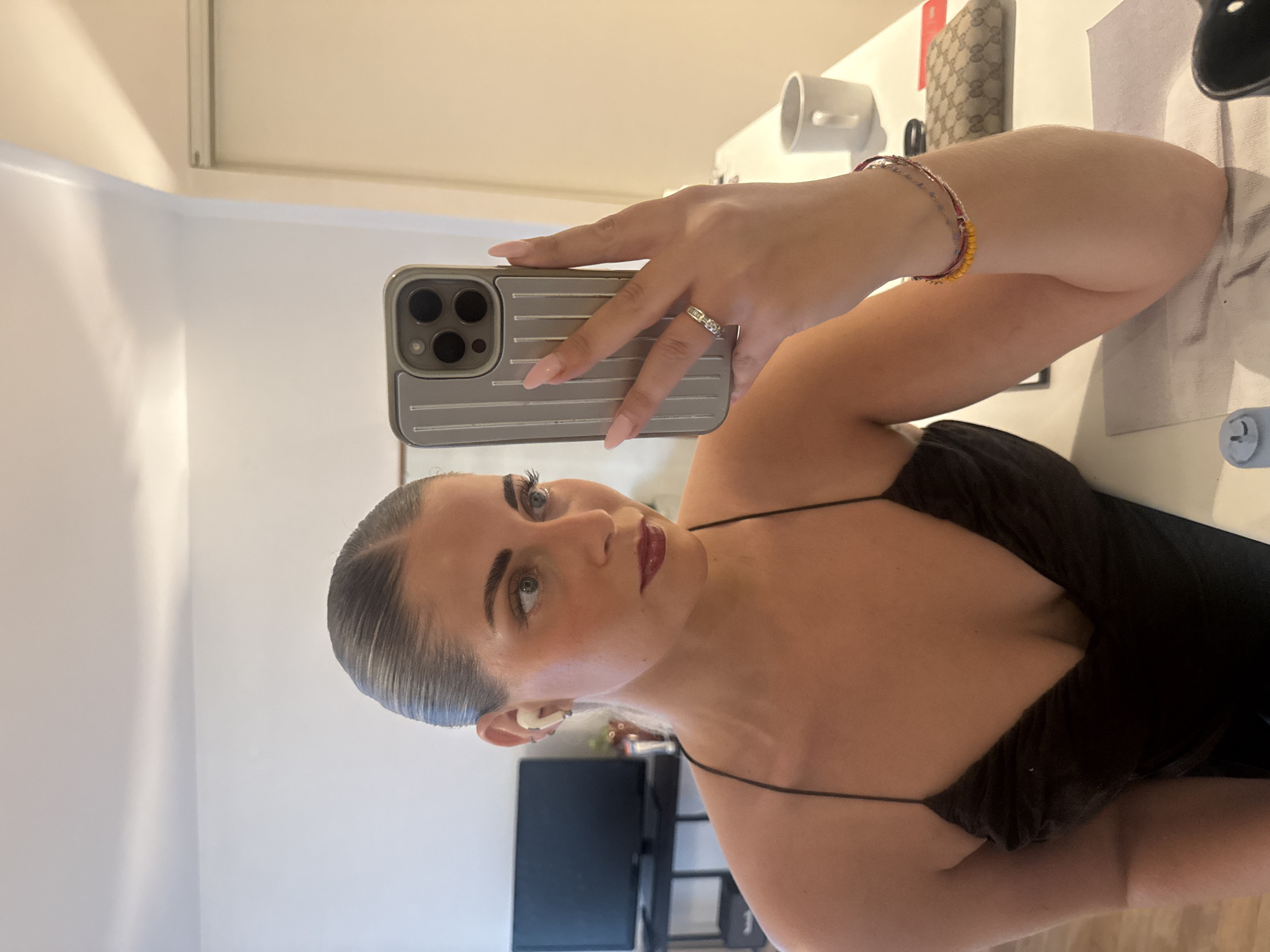 A young woman with her hair slicked back takes a selfie in a mirror. She is wearing a dark, spaghetti-strap dress and has on a ring with three stones on her right hand, and two bracelets on her left wrist. She is wearing AirPods in her right ear. In the background, a television is visible on a stand, and on a table in front of her is a white mug, a wallet, and a red card. The scene appears to be indoors, in a domestic setting. There is no indication that this image was taken in Paris, France.