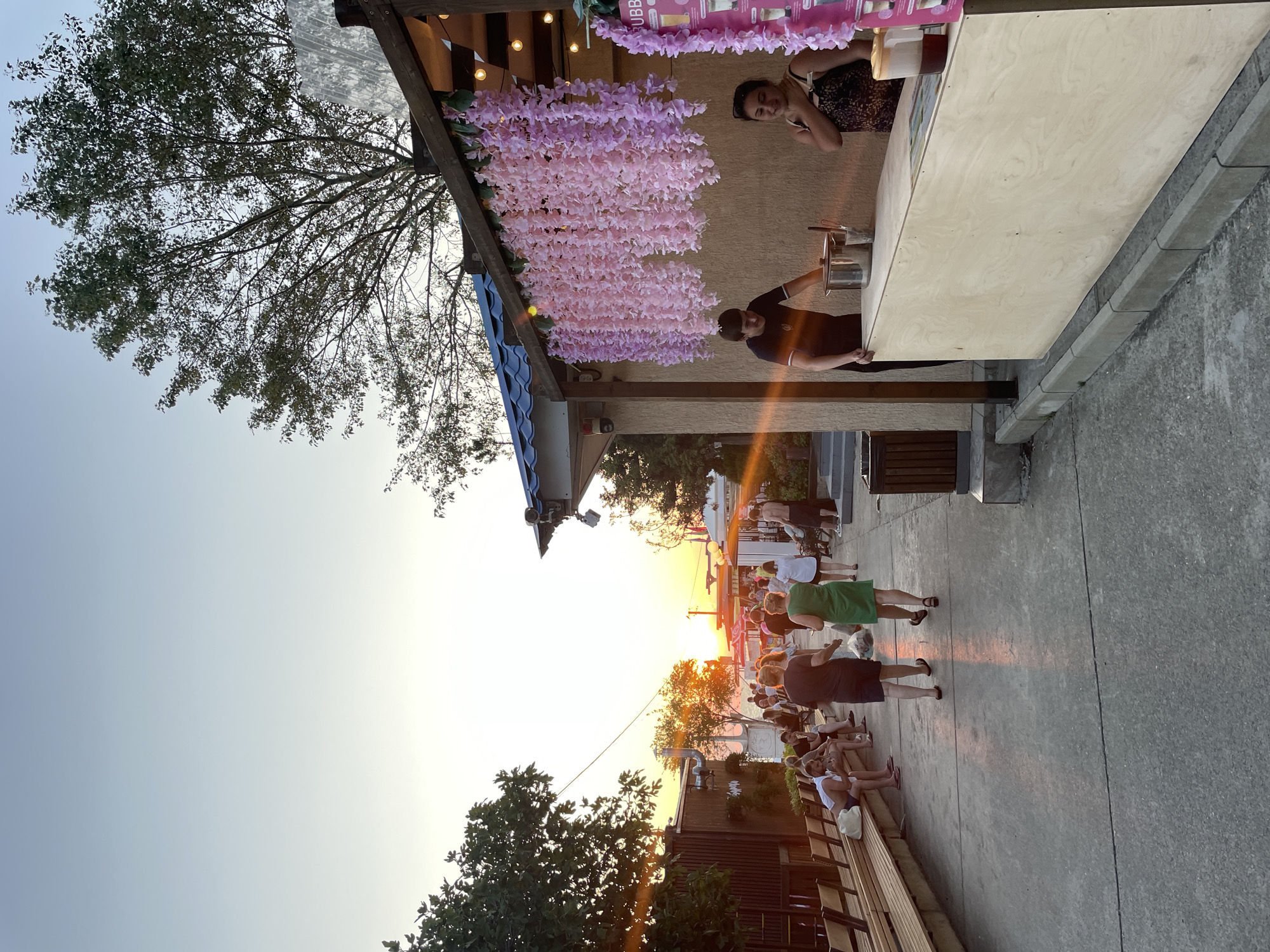 This is a medium shot of a pedestrian promenade in Sochi, Russia, during sunset. The sun is low on the horizon, casting a warm, orange glow across the scene. In the foreground, a man and a woman stand behind a wooden counter at a beverage stand, with pink floral decorations hanging above. The man is preparing a drink, while the woman looks on. A few customers are visible in the background, walking along the promenade or sitting on benches. Trees line the left side of the promenade, and buildings can be seen in the distance. The overall atmosphere is relaxed and casual, with people enjoying the evening at the seaside resort.