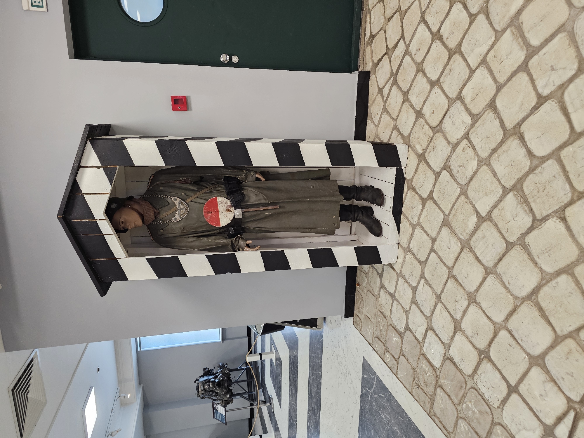 The image depicts a mannequin dressed in a military uniform inside a black and white striped sentry box. The mannequin is wearing a long olive green coat, black boots, and a pilot's helmet. A red and white round object is attached to the front of the coat. The sentry box is positioned in a room with a gray wall and a dark green door above it. The floor is covered with light-colored, patterned tiles. In the background, there is a view of another part of the building containing an airplane engine on a stand. Based on the location provided, it's likely that this scene is located at the Arkhangelskoye Estate Museum in the Krasnogorsky district of Russia.