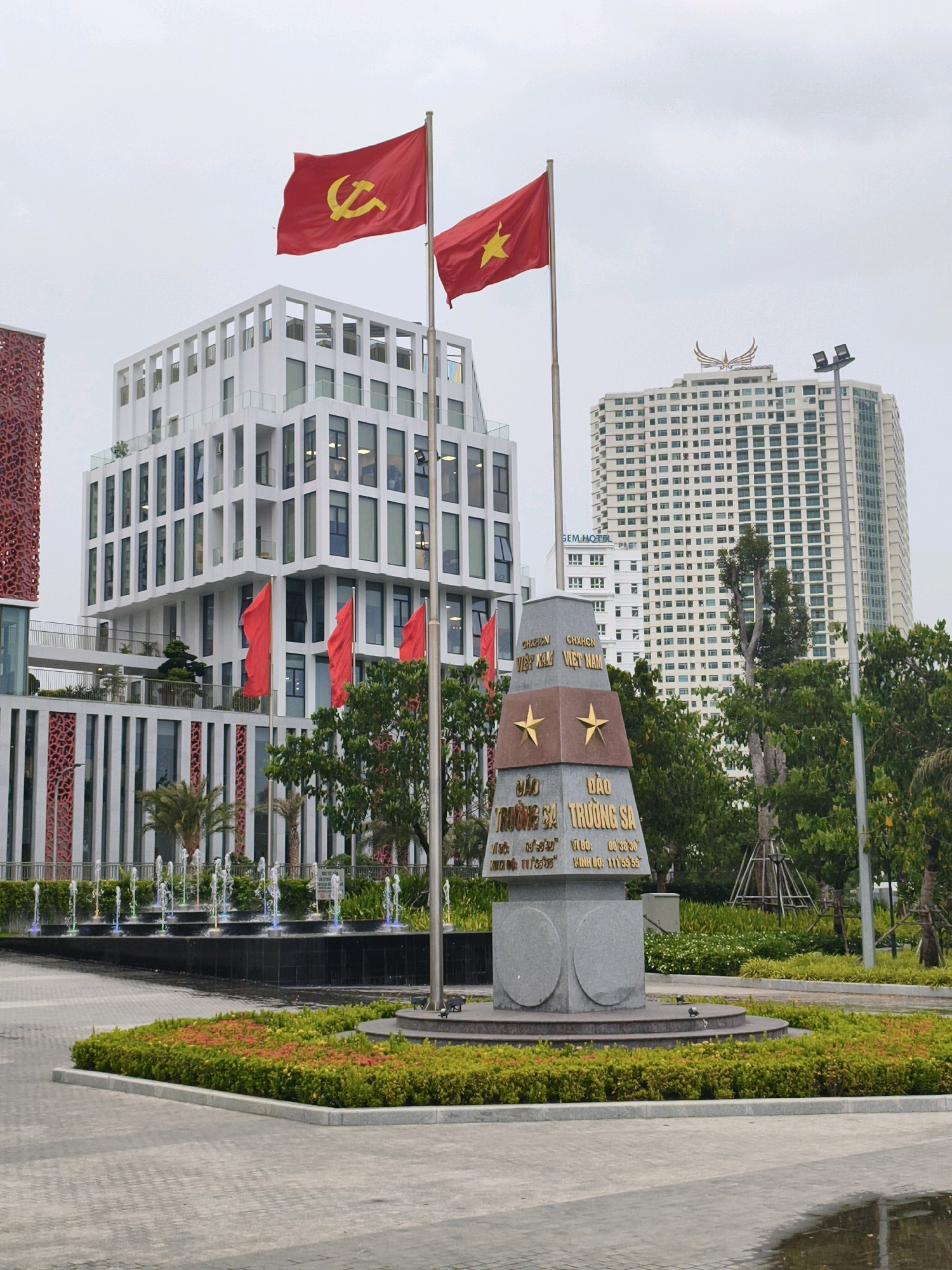 This is a medium-shot, eye-level, outdoor, daytime photograph of a monument and flags in front of modern buildings in Nha Trang, Vietnam. The monument, a conical structure made of stone, is inscribed with Vietnamese text and features two gold stars. Two flags are flying from flagpoles to the left of the monument; the left flag is red with a yellow hammer and sickle symbol, and the right flag is red with a yellow star. Several red flags are also visible on flagpoles to the left of the monument, in front of a white, multi-story building with a patterned facade. In the background, two tall white buildings dominate the skyline, one with a winged crest on its roof. A fountain with colored lights is in the foreground, and manicured greenery and trees surround the monument and fountain area. The ground is paved with light gray tiles. The sky is overcast.