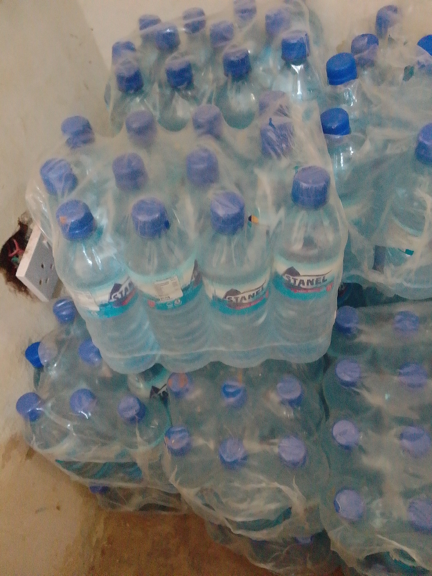 The image shows a pile of bottled water, likely packaged in plastic, with blue caps. The bottles are arranged in multiple rows and columns, suggesting a bulk quantity. The lighting is dim, and the background is indistinct, appearing to be a wall or floor in an indoor setting. A power socket is visible on the wall to the left, partially obscured by some dark material, possibly debris or fabric. The overall impression is that of a storage area. The presence of the power socket and the general appearance of the environment do not specifically indicate a location in Jos, Nigeria, or any other particular city.