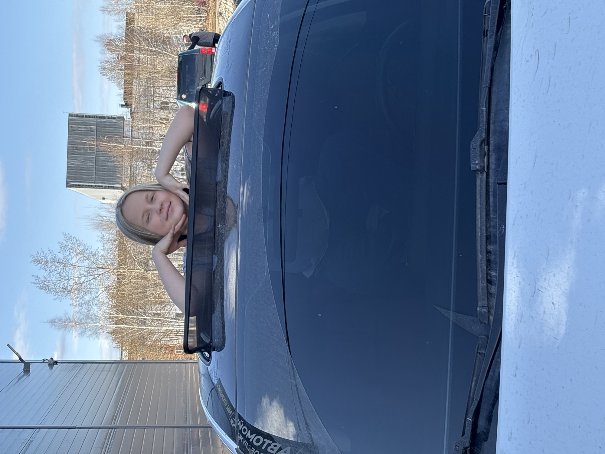 A young girl with light blonde hair is looking out of the open window of a black car. She is resting her face on her hands. The scene is outdoors, with a building to the left, trees and other cars in the background. The location appears to be Izhevsk, Russia, based on the mirrored "BLOWOMOTRA" sticker on the car, which when reversed, reads "Автомойка" (Car Wash in Russian), and a possible local business name "ВПИ ОАО ИЖСПУТНИК" (VPI OAO IZHSputnik). The day is bright and sunny.