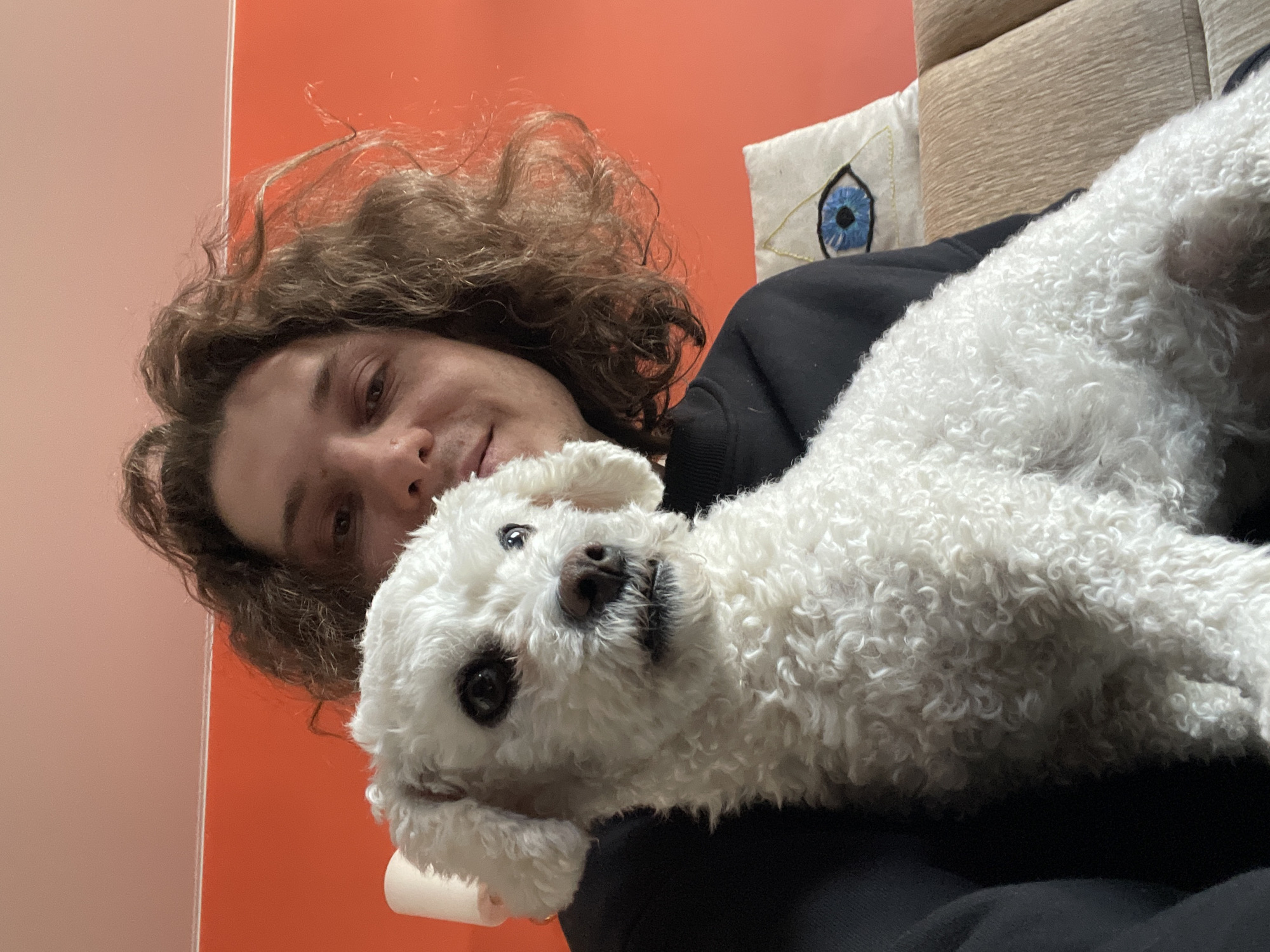 A person with curly brown hair is holding a white Bichon Frise dog. The person is wearing a black hoodie. The background includes an orange wall and a beige pillow with an embroidered blue eye. The location is not discernible from the image and therefore cannot be determined to be in Yekaterinburg, Russia.