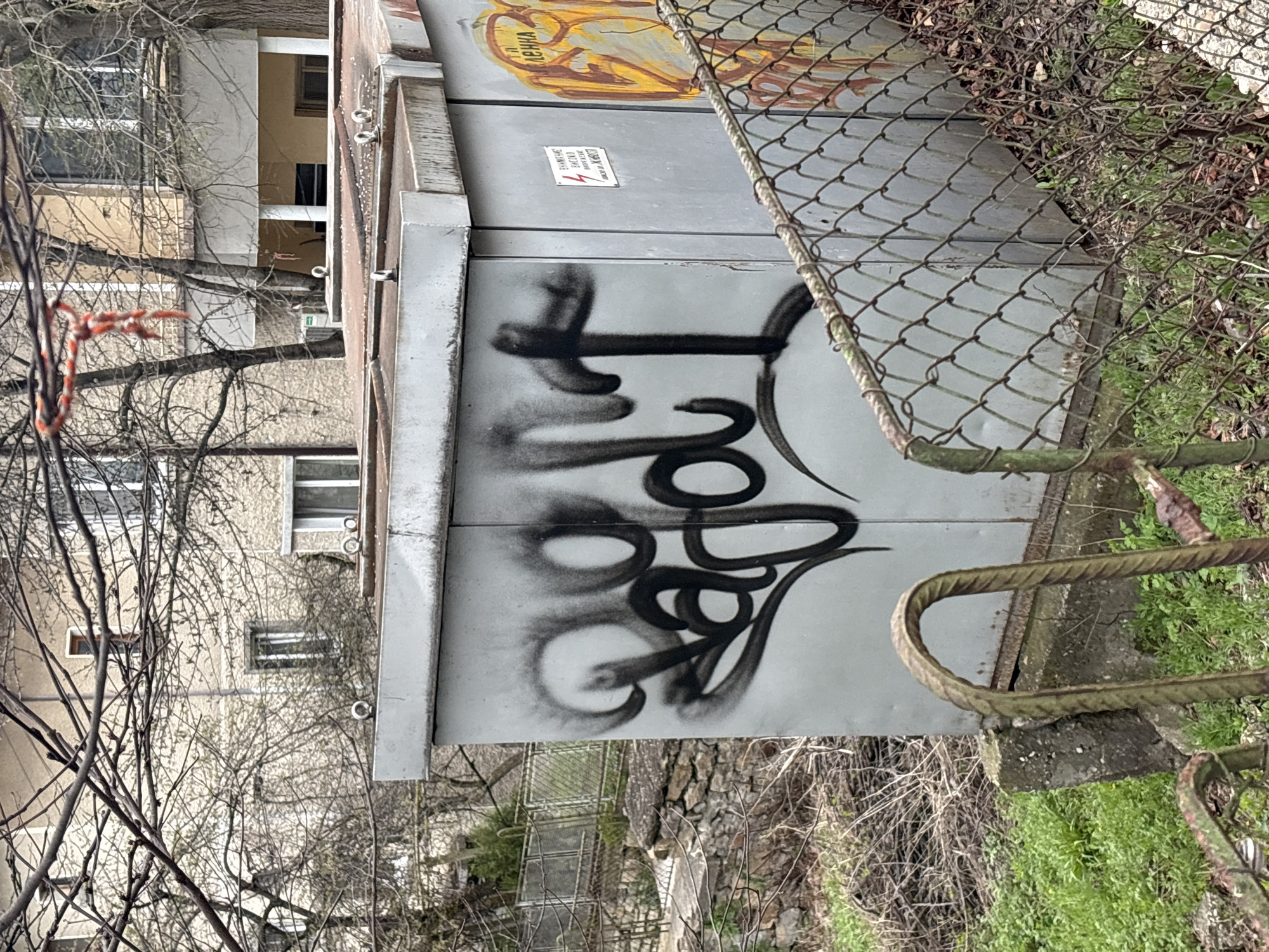 A ground-level, medium shot depicts a graffitied utility box in Veliko Tarnovo, Bulgaria. The box, painted a flat gray, is adorned with illegible, stylized black graffiti. Above the graffiti, there's a warning sign written in what appears to be Cyrillic script. A crude, yellow and orange painting is visible on the upper section.
To the right of the box, a rusted chain-link fence runs along a drop-off to an area overgrown with grass and small vegetation. A rusted metal bar is present in the lower right corner.
The background shows a multi-story building with tan and white facade and visible windows, partially obscured by bare tree branches. More bare branches are visible in the lower left corner of the frame. A red and white cable is also visible on the left side.