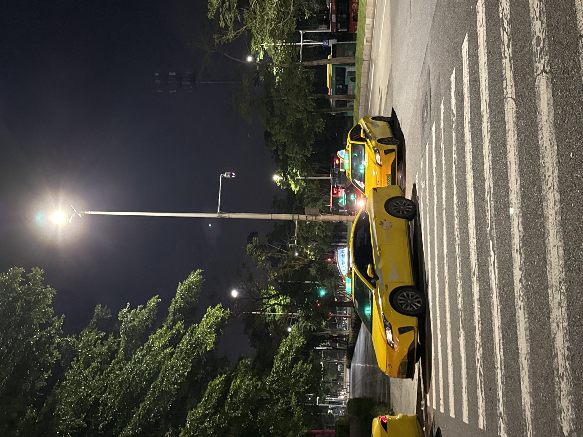 This image shows a street scene at night in Haizhu District, China. Two yellow taxis are parked on the right side of the road, next to a pedestrian crossing. The taxi in the foreground is facing towards the camera, while the taxi behind it is partially visible. The street is wet, suggesting recent rain. Tall trees line the left side of the road, their leaves illuminated by streetlights. In the background, there are more trees and buildings, as well as a red and green traffic light. The sky is dark, indicating it is nighttime. There is some text visible on the side of the taxi in the foreground, but it is too blurry to read. The overall mood of the image is quiet and slightly mysterious, with the glowing lights of the city contrasting with the dark night sky.