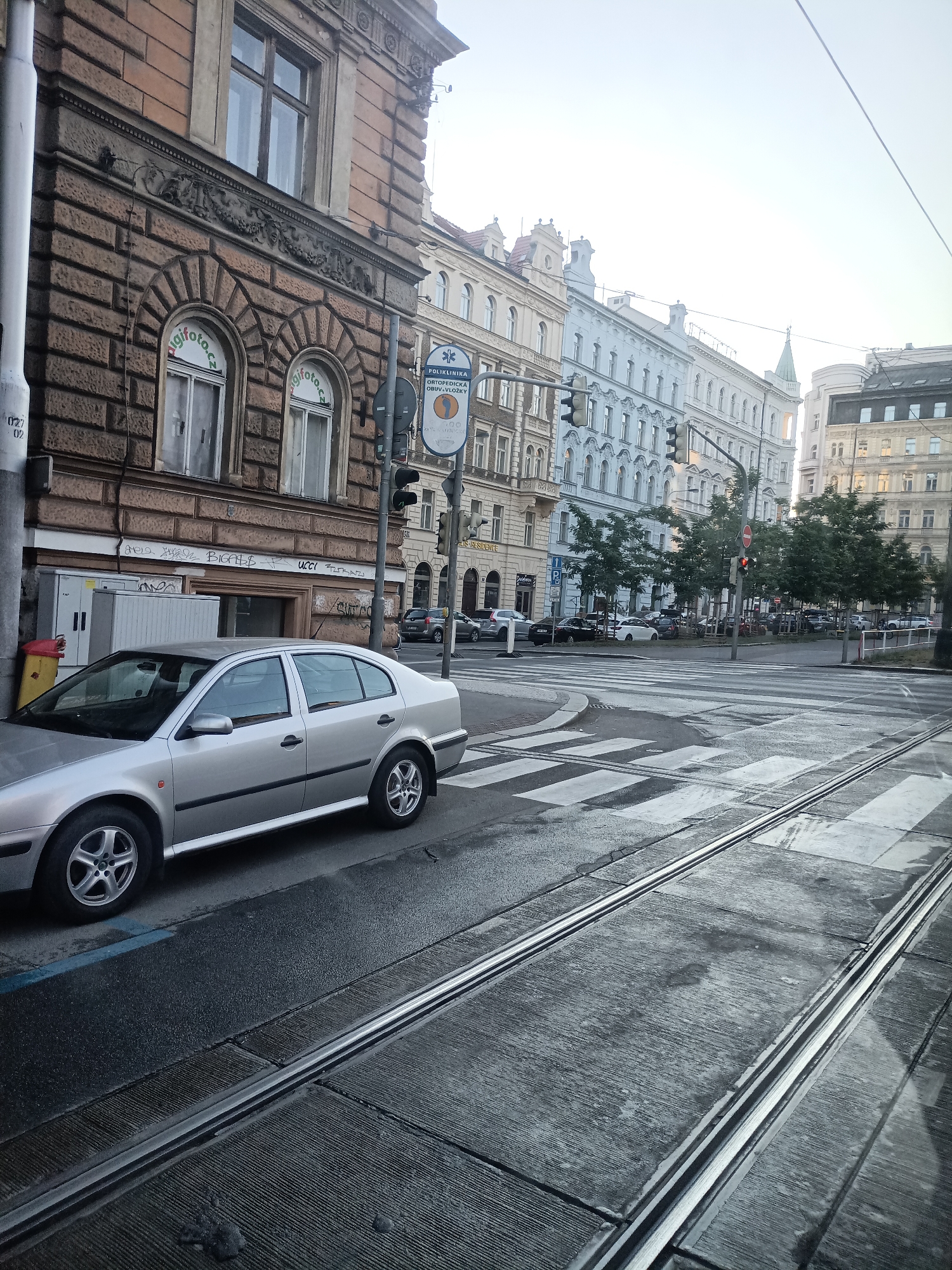 A silver sedan is parked on a wet street next to tram tracks. The sedan is in the foreground, angled to the left. Behind the car, buildings line the street, featuring classic architecture with intricate stone carvings and arched windows. One building has a sign for "poliklinika ortopedicka obuv" which translates to "orthopedic shoe polyclinic." The street appears to be an intersection with a pedestrian crosswalk visible. Traffic lights are present, with one showing a red light. Several trees are planted along the sidewalk. The sky is overcast and bright. The tram tracks are in the lower half of the image, running parallel to the car and extending into the distance. The overall impression is an urban street scene in Prague, Czechia.