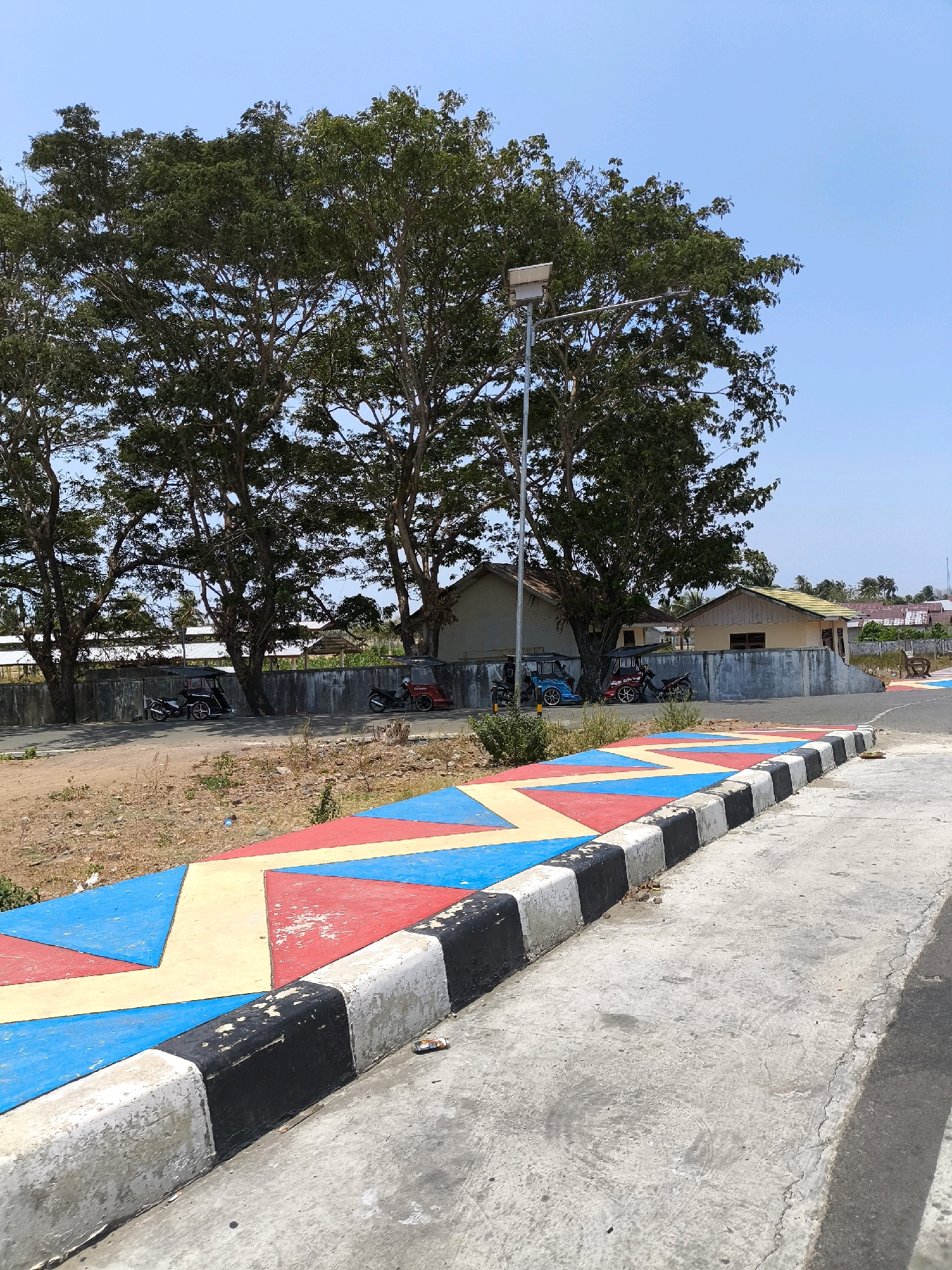 The image depicts a bright, sunny day scene in Kwandang, Indonesia. In the foreground, a paved pathway with a decorative, brightly colored border of red, yellow, and blue geometric shapes runs along a gray concrete curb. The curb is painted with alternating black and white stripes, some of which show signs of wear. Beyond the curb, a patch of dry, uneven ground is visible with sparse vegetation.
In the middle ground, a low concrete wall runs horizontally across the image. Behind the wall, several buildings with light-colored walls and yellowish roofs are situated. A tall street lamp stands near the center of the image, its light fixture illuminated, positioned amidst a cluster of large, leafy green trees. Parked alongside the concrete wall are several motorcycles, including two three-wheeled vehicles that appear to be motorized rickshaws or "bajaj". Two individuals are visible standing near these vehicles.
The background reveals more buildings, palm trees, and a clear blue sky. The overall impression is that of a semi-urban or suburban area, likely a public space or roadside area within Kwandang.