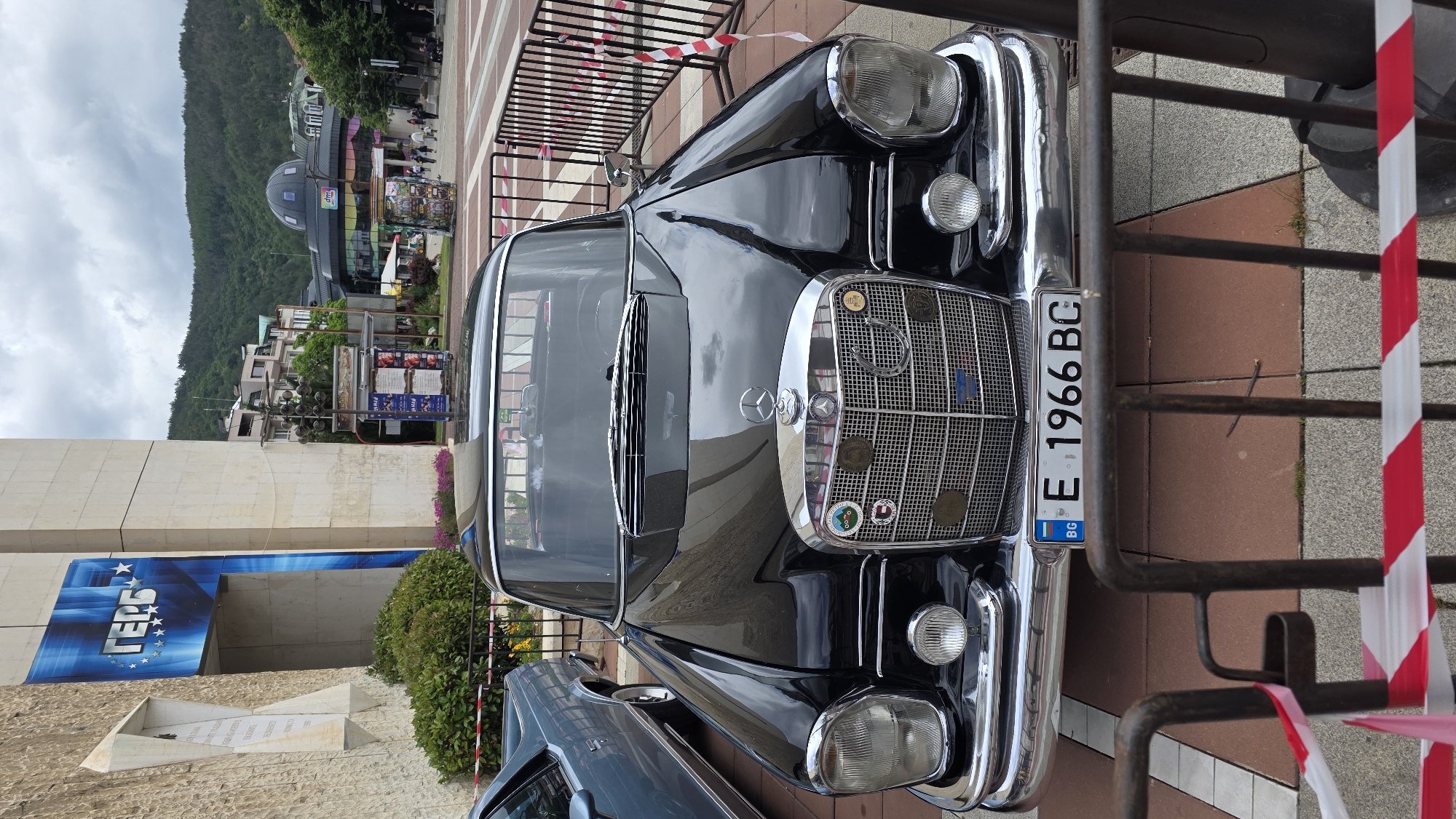The image captures a vintage black Mercedes-Benz, license plate E 1966 BC, parked in Blagoevgrad, Bulgaria. The car is positioned in front of the American University in Bulgaria's building. Part of a fenced pedestrian area surrounds the vehicle. The background includes buildings, greenery, and a hillside. The sky is overcast.