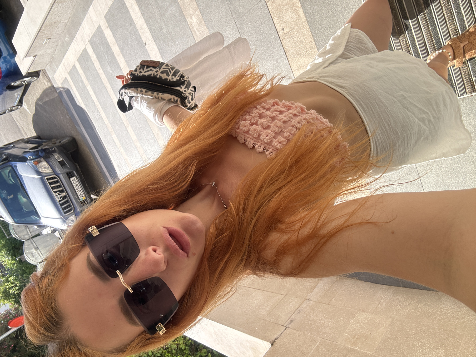 A woman with ginger hair is taking a selfie outdoors. She is wearing large, dark sunglasses, a pink crocheted top, and a white skirt. She is holding a patterned bag in her right hand. In the background, a dark blue SUV is parked. The scene appears to be in a city, possibly Budva, Montenegro, with paved walkways and greenery visible. The lighting suggests it is a sunny day.