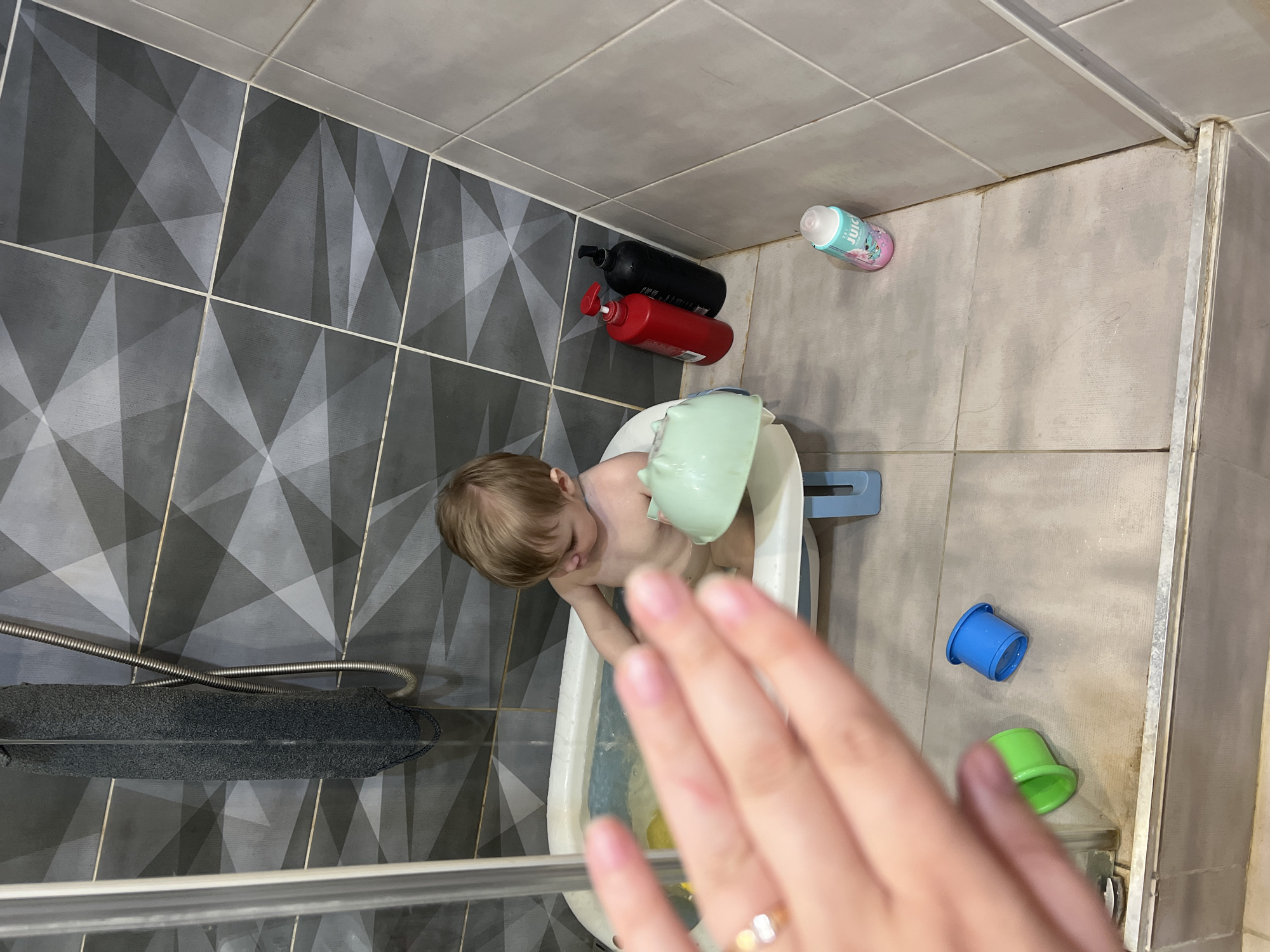 A child with blond hair is in a white plastic bathtub, playing with a green toy watering can. The child is partially obscured by a person's hand with a ring on the ring finger. The bathroom has gray and white patterned tiles on one wall and solid light gray tiles on another. There are bottles of shampoo and lotion near the wall, and plastic cups near the tub. A gray bath sponge hangs next to the shower head. The scene is in the city of Noviy, Russia.