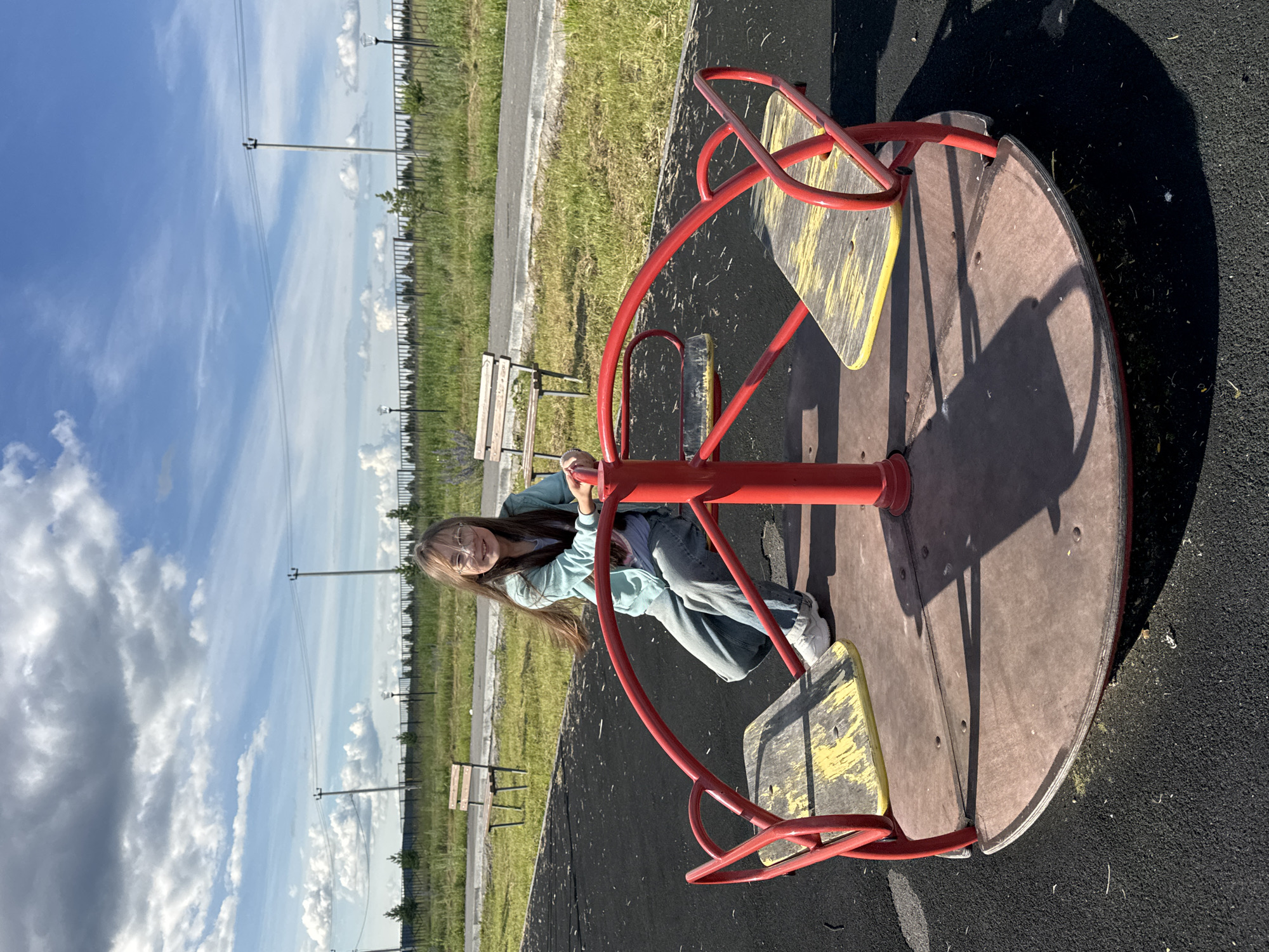 A young woman with long brown hair and glasses is seated on a merry-go-round in a park. She is wearing a light blue hoodie and blue jeans. Her left hand is gripping the red metal bar of the merry-go-round, and she is smiling at the camera. The merry-go-round has red metal poles and brown seats with yellow accents. It is situated on a dark grey, rubberized surface, typical of a playground.
In the background, there is a strip of green grass bordered by a black fence. Beyond the fence, more grass and some trees are visible, along with several lampposts. The sky is a vibrant blue with scattered white clouds. The image appears to be taken in the daytime, likely late afternoon given the shadows cast by the merry-go-round and the lampposts. The weather seems pleasant, with clear skies and sunshine. The scene suggests a casual outing or leisure time at a playground in Mursalimkino, Russia. There is no visible text in the image.