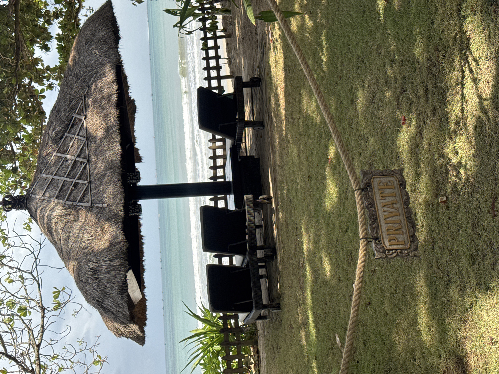 The image depicts a tranquil scene at a beachside resort in Kuta Selatan, Indonesia. In the foreground, a thick, weathered rope barrier separates a manicured lawn from the beach. A decorative "PRIVATE" sign hangs from the rope, indicating exclusivity. Behind the rope, two dark-colored lounge chairs are neatly arranged on the grass, facing the ocean. Further back, beneath a large, thatched-roof umbrella, three more lounge chairs are positioned on the sandy beach, providing ample seating for relaxation. The ocean stretches out to the horizon, its turquoise waters gently lapping against the shore. A light breeze rustles the leaves of the trees overhead, casting dappled shadows on the scene. The overall impression is one of serene seclusion and luxurious comfort.