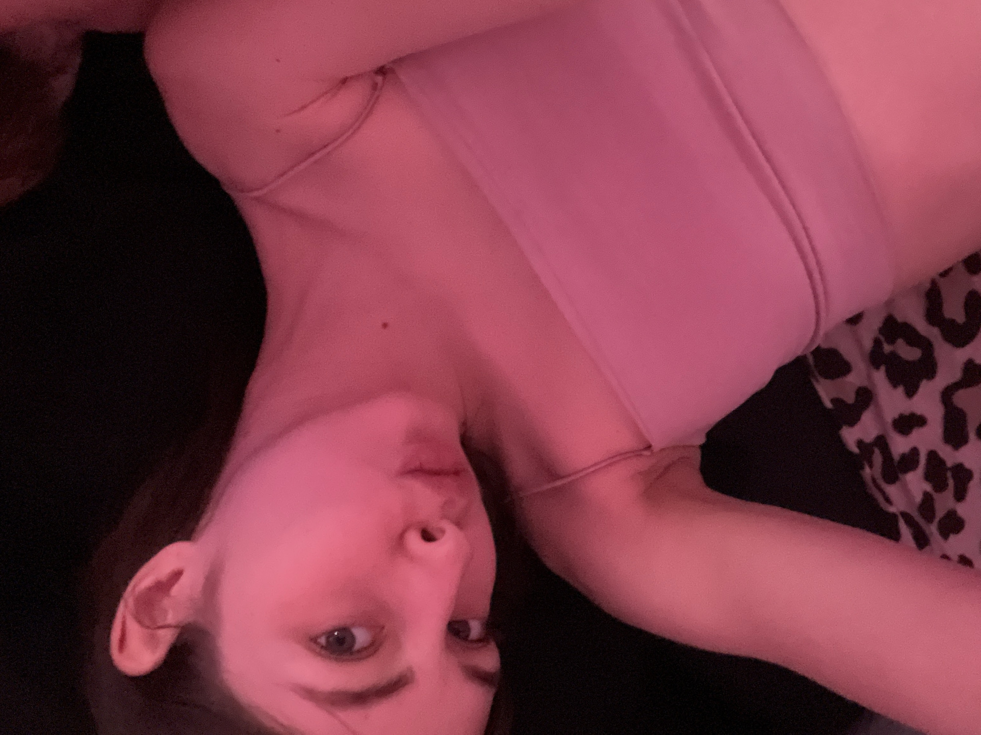 A woman is lying down, wearing a pink sports bra. She is looking at the camera. The background includes a black surface and a black and white animal print fabric. The scene is lit with a pink light. There is no indication of the location being in Saint Petersburg, Russia.