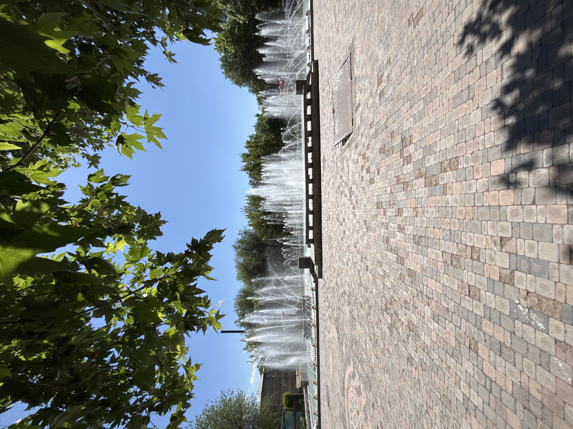The image depicts a public plaza in Yerevan, Armenia, featuring a large, multi-tiered fountain. The fountain is in operation, with numerous jets of water spraying upwards. The plaza is paved with interlocking bricks in various shades of brown, beige, and gray. Green trees and foliage frame the top of the image, casting dappled shadows on the ground. In the background, more trees and some buildings are visible under a clear blue sky. There are no people visible in the image. A metal manhole cover is present in the foreground. The overall impression is one of a well-maintained, sunny public space designed for relaxation and recreation.