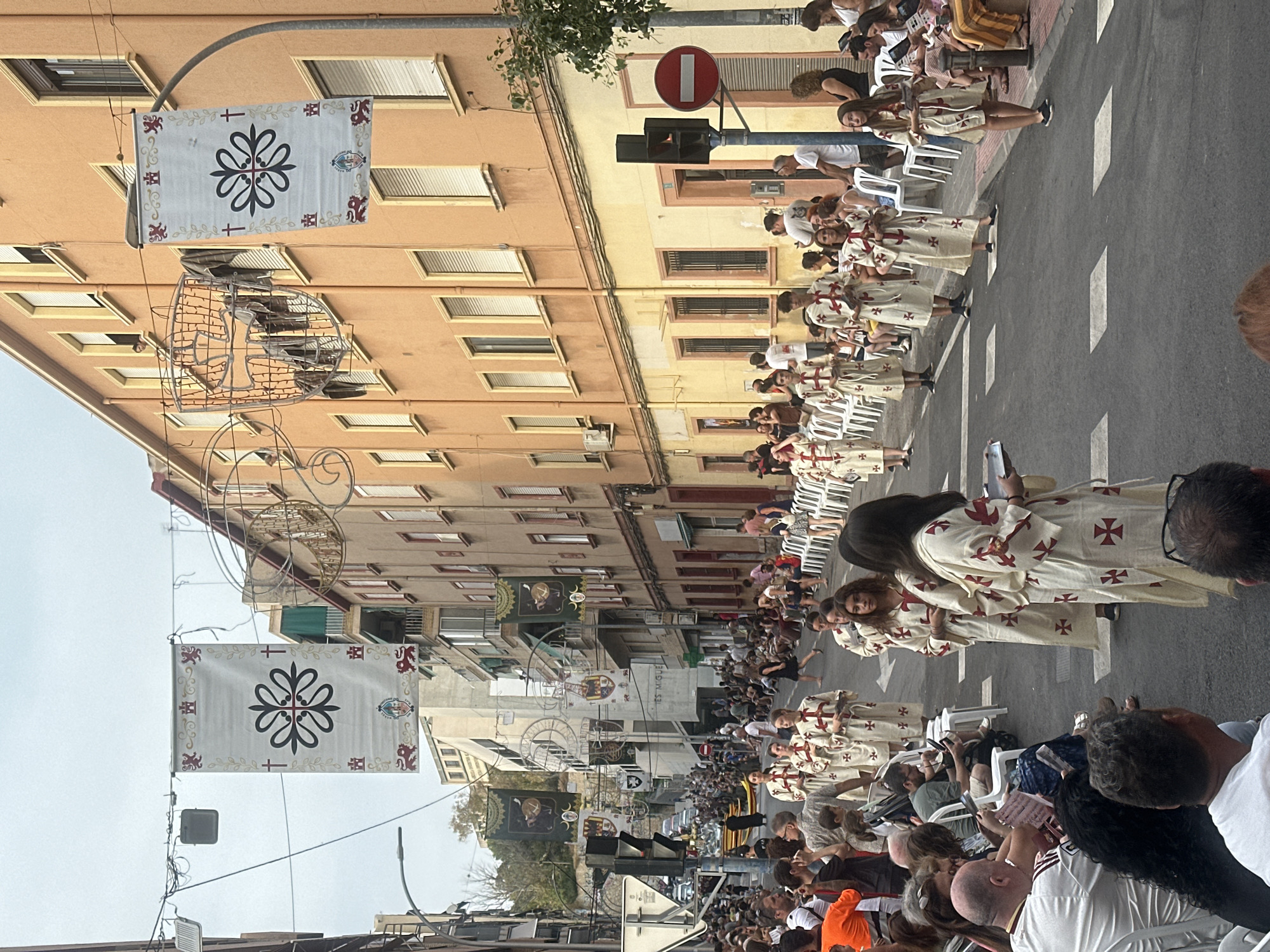 This is a medium-shot, eye-level, outdoor photograph of a street procession in Alicante, Spain. The procession appears to be a religious or cultural event, with participants dressed in ceremonial robes. Several people are walking down the street, some holding what appear to be religious artifacts or scrolls. They are wearing long, white robes adorned with red cross symbols. Spectators are seated on white plastic chairs lining the sides of the street, watching the procession. Buildings with balconies and shuttered windows line both sides of the street. Several banners with a floral design and religious symbols are hung across the street overhead. Traffic lights and a red circular "no entry" sign are visible. The sky is overcast.