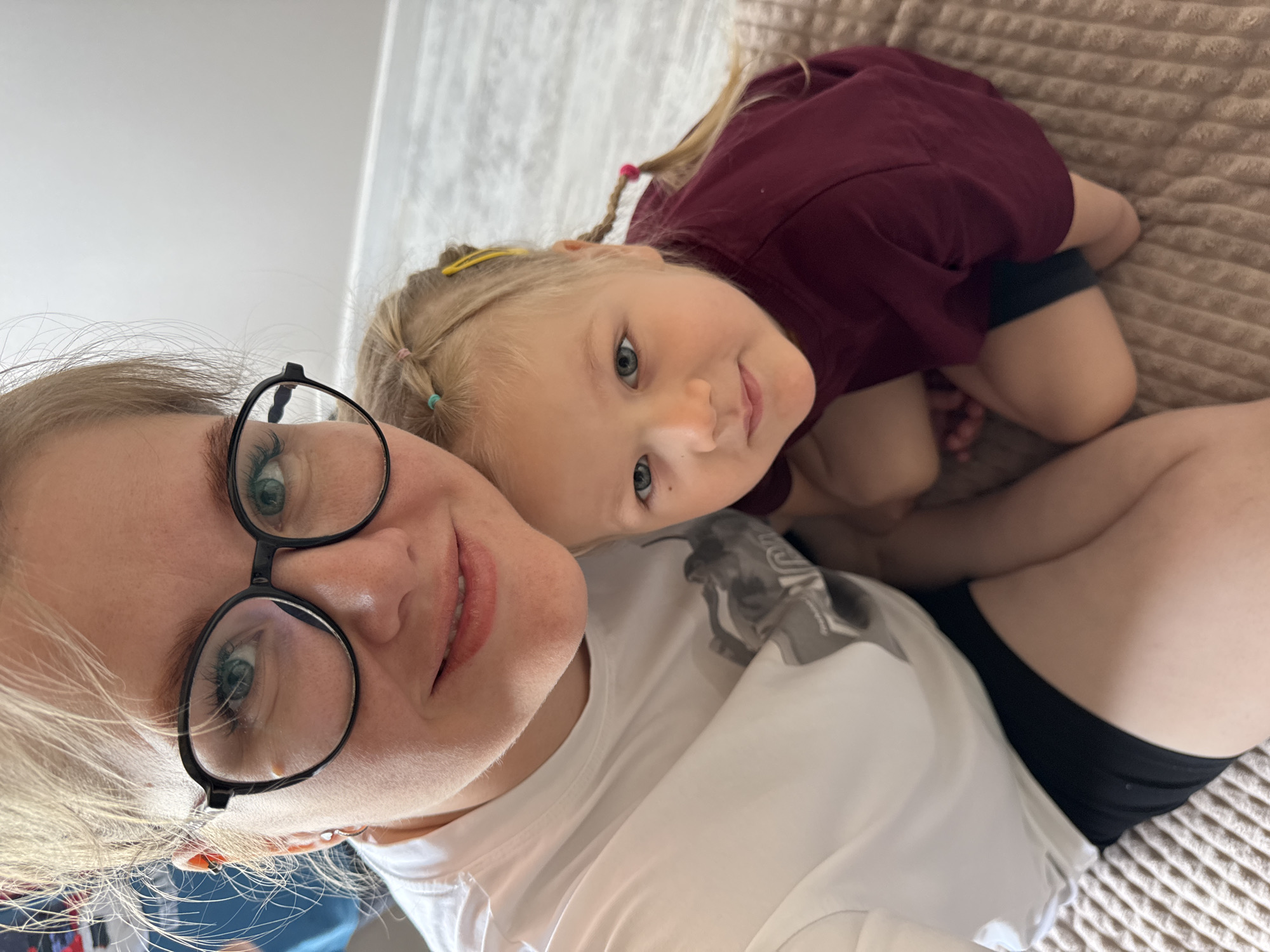 A photograph shows a woman and a young girl lying on a beige surface, possibly a bed. The woman, positioned closer to the camera, wears glasses and a white t-shirt. The young girl, with blonde hair styled in braids with colorful hair ties, is wearing a maroon t-shirt and dark shorts. The photograph is taken at an angle, with the subjects' faces tilted towards the camera. The photograph does not contain enough information to confirm a location in Gornyi (Dvurechenskoe S/P), Russia.