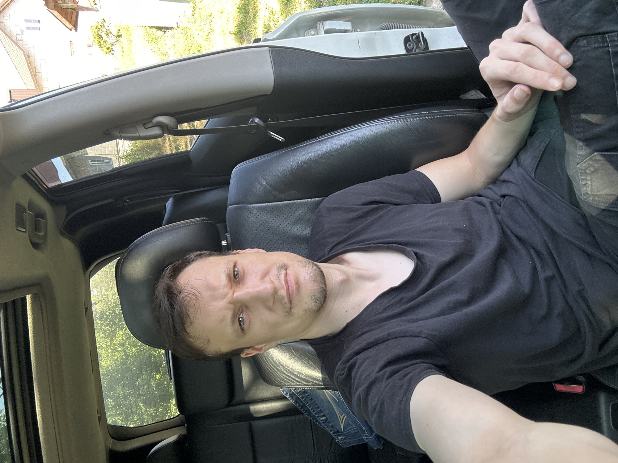 A medium shot photograph shows a man sitting in the driver's seat of a car, facing the camera. The man is wearing a black t-shirt and dark pants. He has short brown hair and stubble. His left arm rests on the seat next to him, and his right hand is resting on his lap. The interior of the car is visible, including the leather seats and a sunroof. The car appears to be parked outdoors, with trees and buildings visible in the background. The overall lighting suggests it is daytime. The scene is located in Makiv, Ukraine.