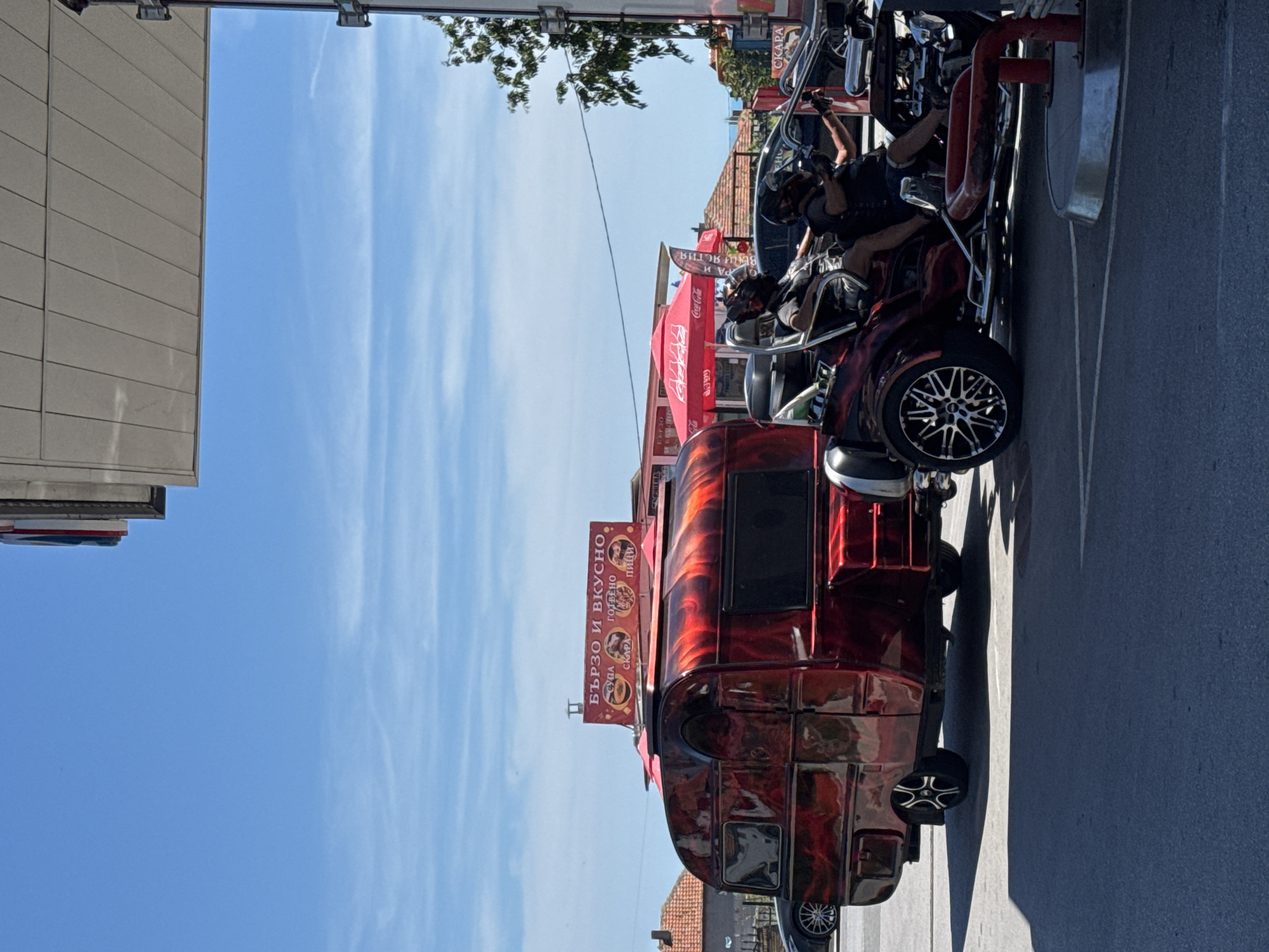 In the city of Byala, Bulgaria, on a bright, sunny day, a custom red and black flame-patterned trailer is parked next to a trike with two occupants wearing helmets. The trailer has a small window on the side and a storage compartment. A red awning with "Coca-Cola" branding is visible behind the trailer. To the left of the trailer, a dark-colored car is parked. Further in the background, several buildings with red tile roofs are visible. The scene appears to be near a gas station, as indicated by the overhead structure and a fuel pump in the foreground. A sign above the trailer reads "БЪРЗО И ВКУСНО" (Fast and Delicious) and lists food items such as "СУПА" (Soup), "СКАРА" (Grill), "ГОТВЕНО" (Cooked), and "ПИЦИ" (Pizzas). The sky is clear blue with a few wispy clouds.