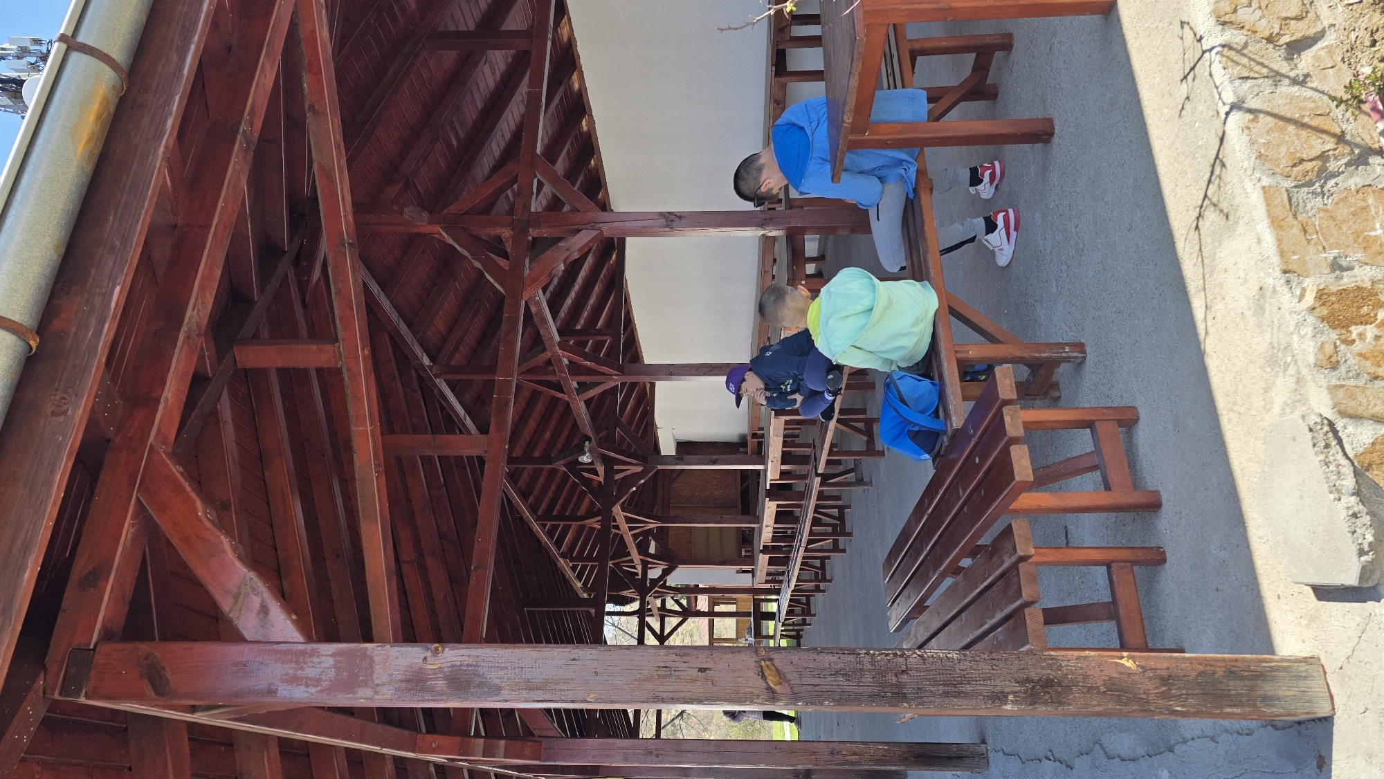 Two boys are seated at a wooden picnic table under a wooden-framed building. The boy on the left wears a light blue jacket and gray pants and the boy on the right wears a blue jacket, dark pants, and a purple hat. They are both looking down. A blue backpack is next to the boy on the right. The building is located in Elenovo, Bulgaria. The building has a dark reddish-brown wooden frame and a roof. The benches are also made of reddish-brown wood. Outside of the building is a concrete floor and a stone wall.
